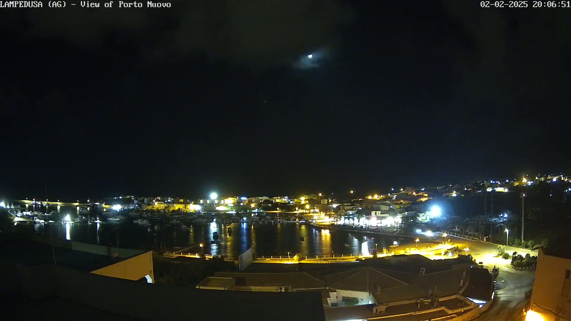 A nighttime view of a harbor town with boats docked, lit up buildings, and a mostly clear sky with a moon visible.