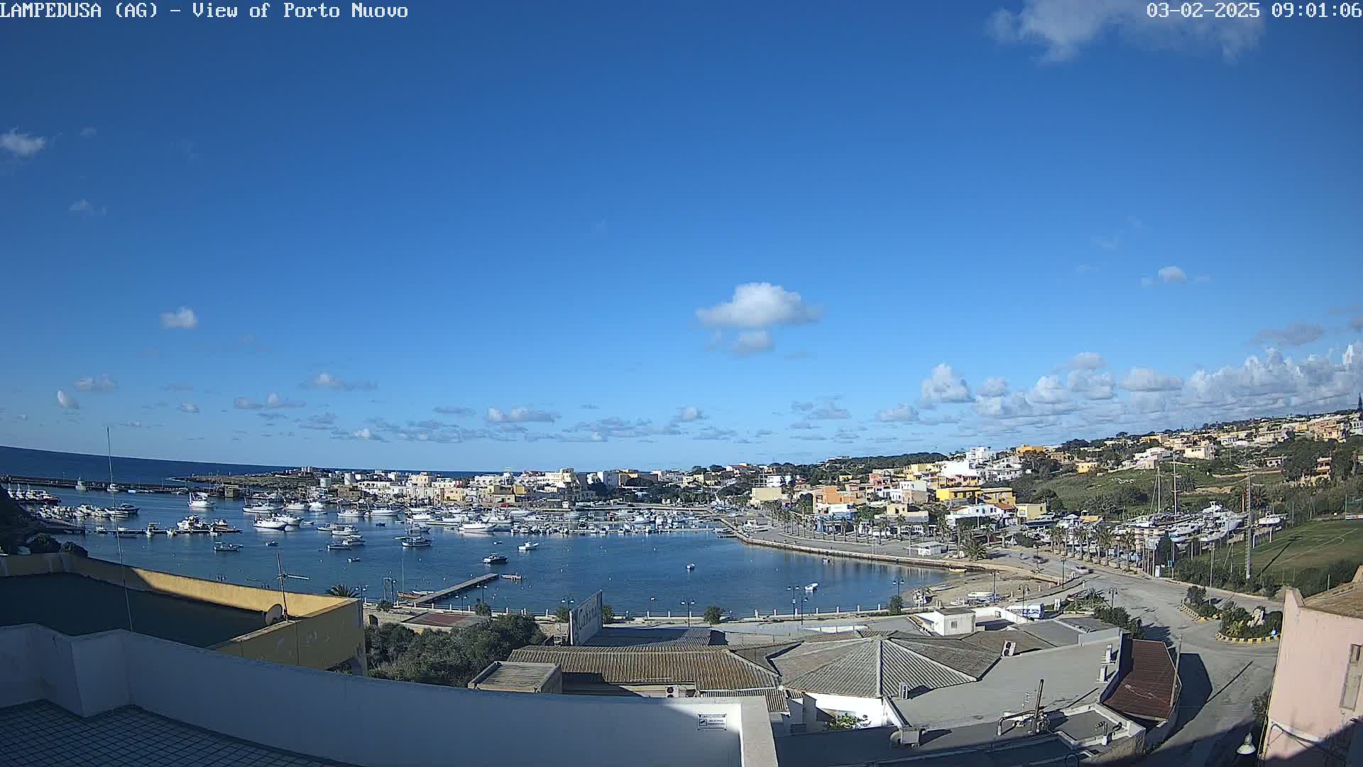 A harbor filled with boats is nestled within a town situated on a coast under a mostly clear blue sky with scattered clouds.