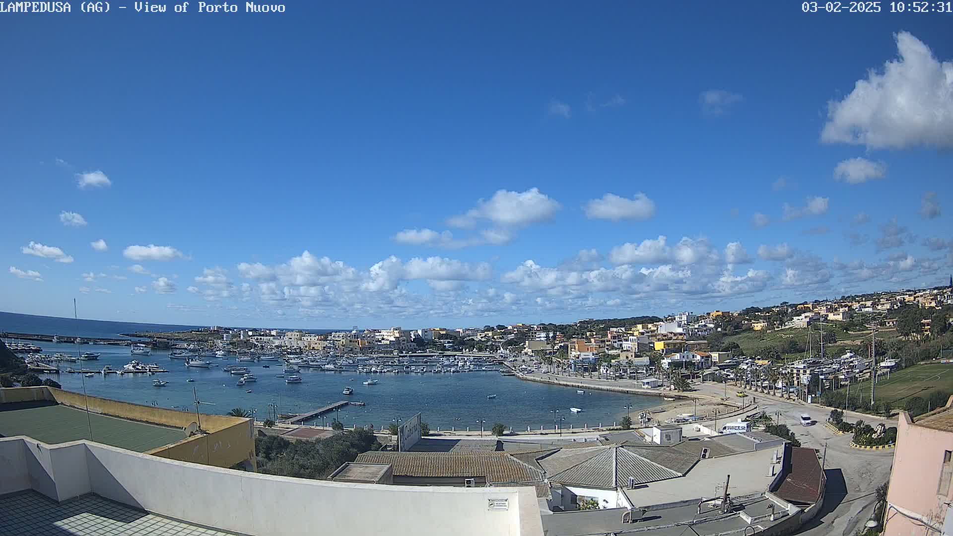 A harbor filled with boats is nestled in a town under a mostly sunny sky with scattered cumulus clouds.