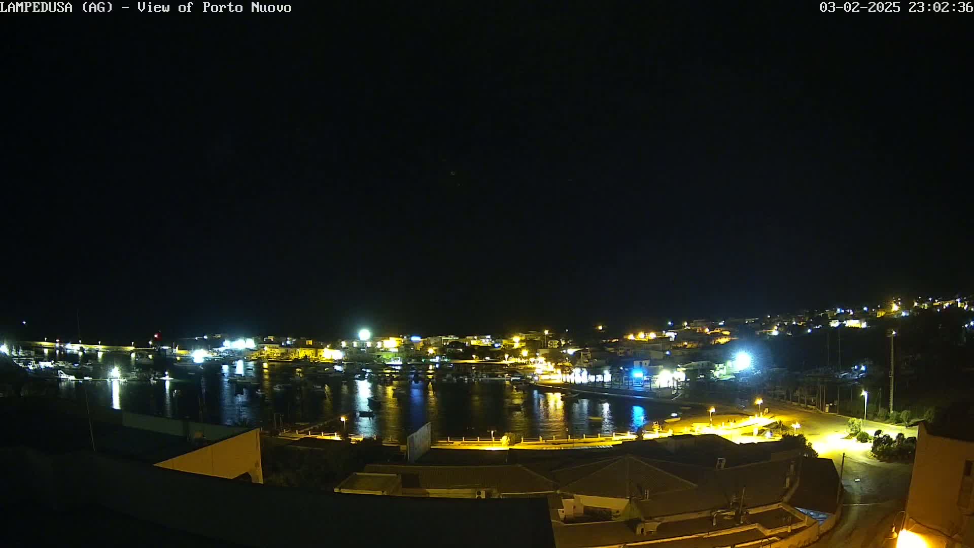 A nighttime, high-angle view of a harbor town with brightly lit buildings and boats in calm water under a clear, dark sky.