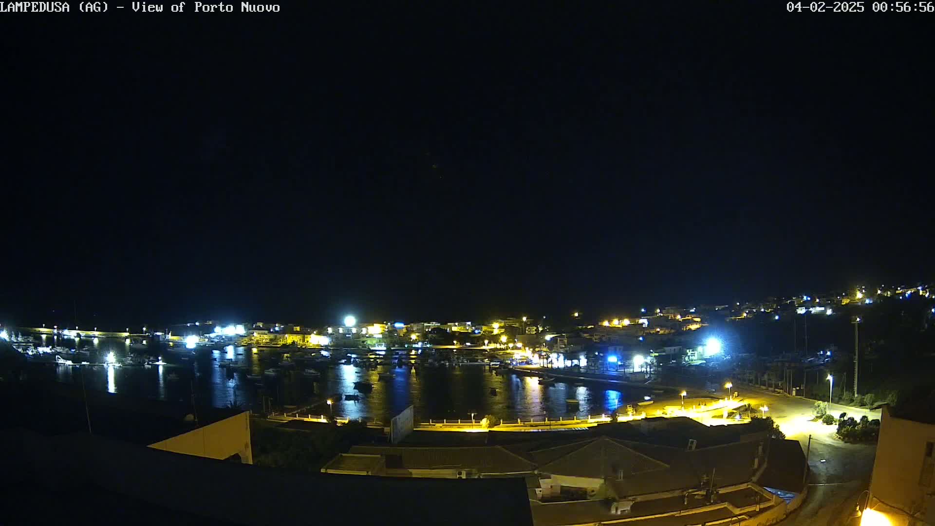 A nighttime, high-angle view of a harbor town with its lights illuminated, reflecting on the calm water.