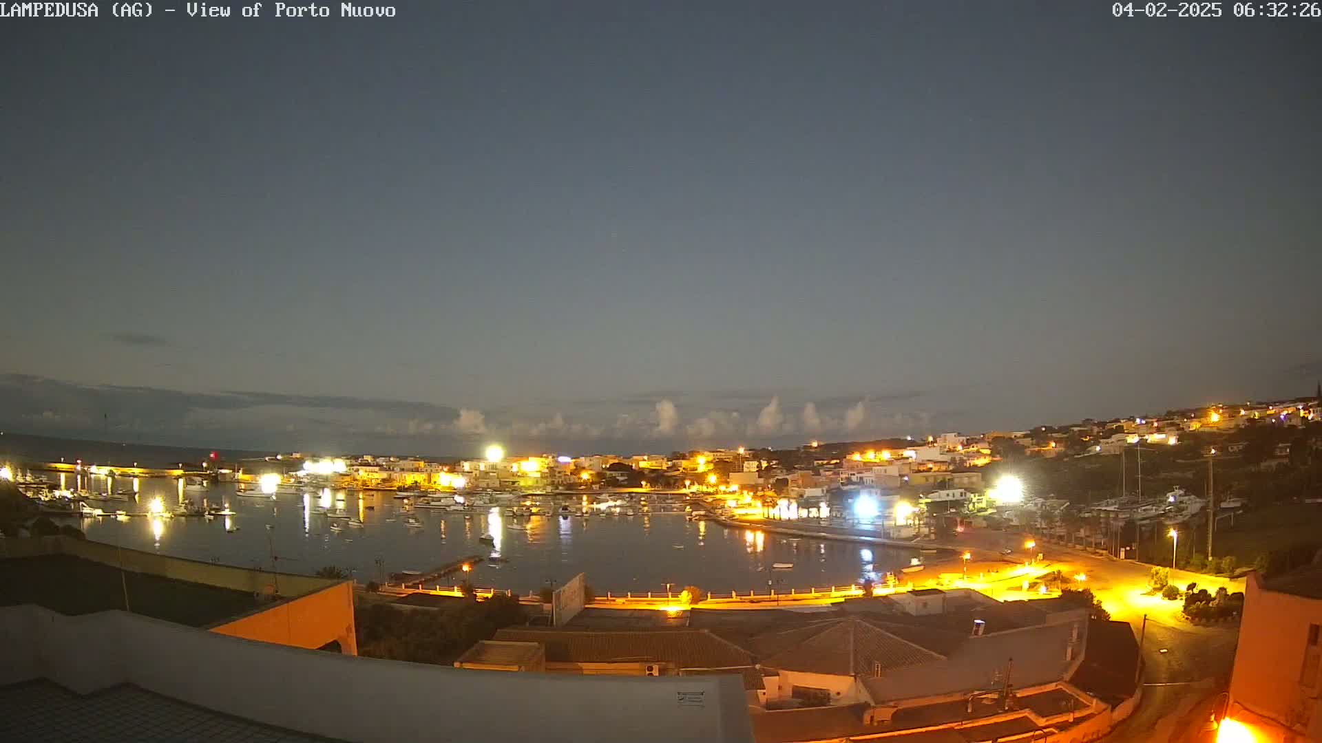 A nighttime view of a harbor town with boats in the water, illuminated buildings, and a mostly clear sky.