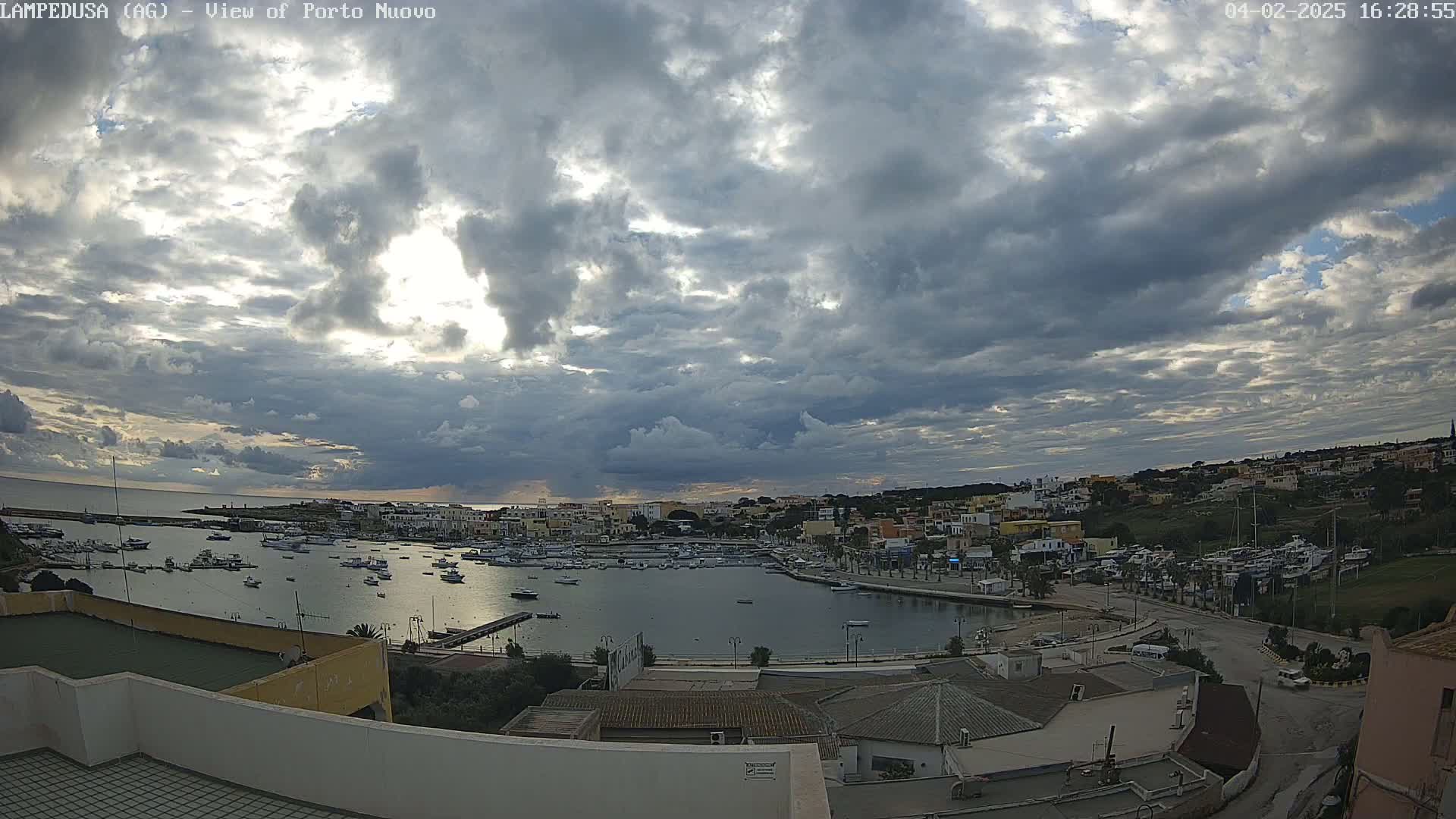 A harbor filled with boats is surrounded by buildings under a mostly cloudy sky with some sunlight breaking through.