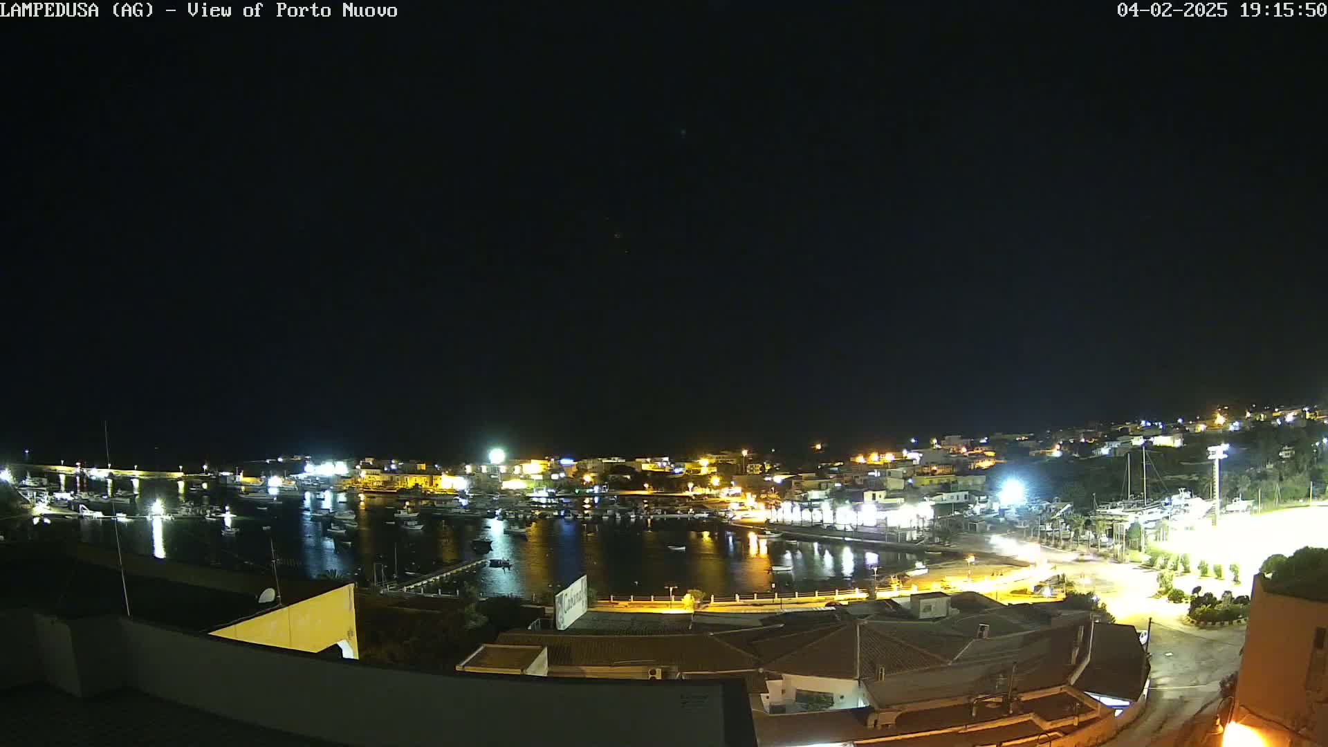 A nighttime, aerial view of a harbor filled with boats, surrounded by a town with illuminated buildings under a clear, dark sky.