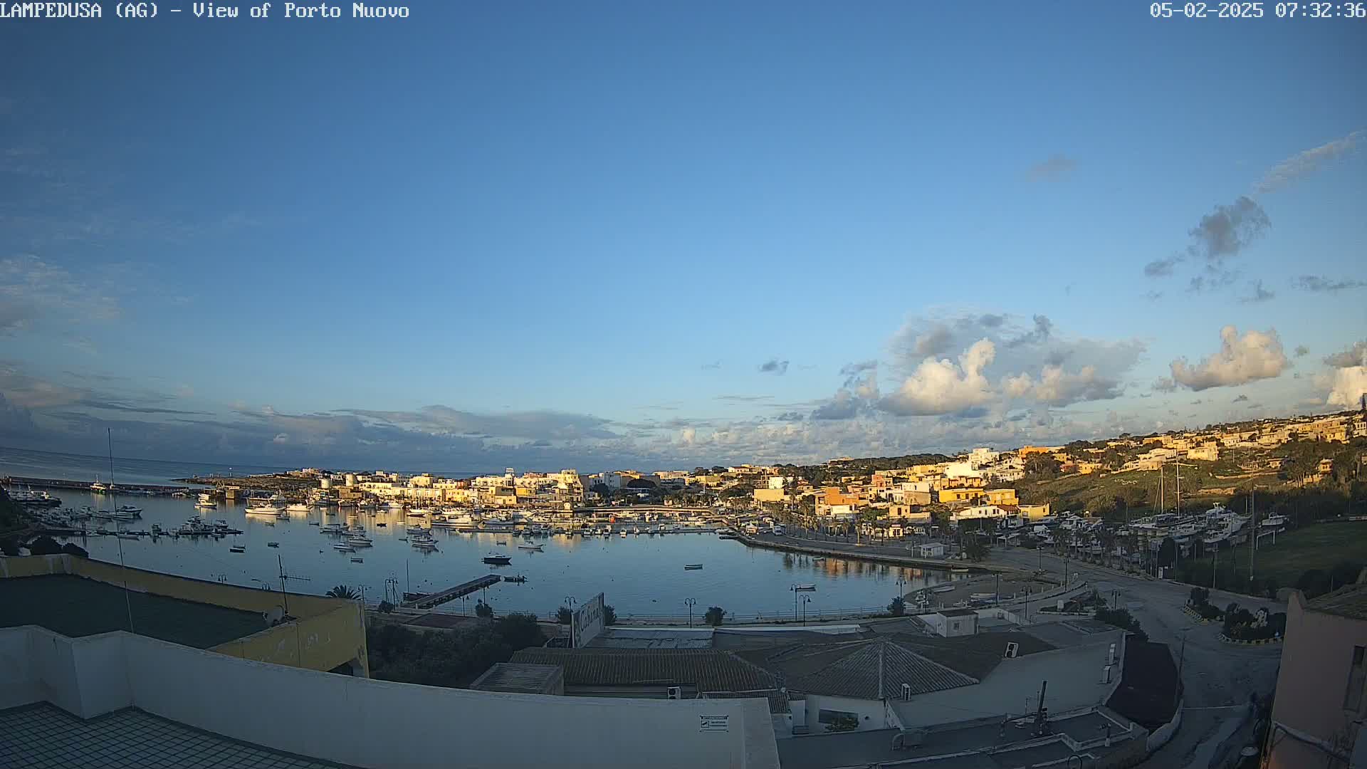 A harbor filled with boats is nestled in a town of low-rise buildings under a mostly clear sky with scattered cumulus clouds.