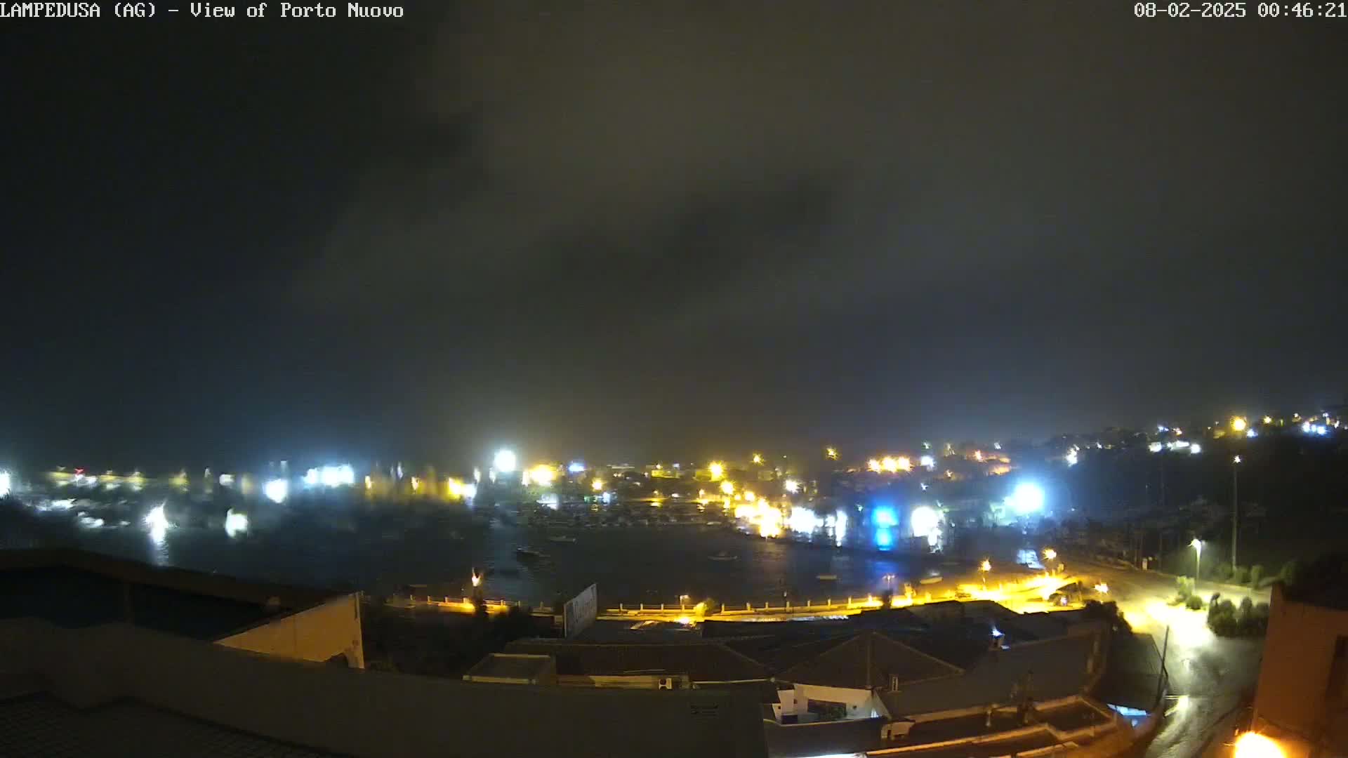A nighttime view of a harbor town under a dark, cloudy sky with rain, showing brightly lit buildings and a partially obscured harbor with boats.
