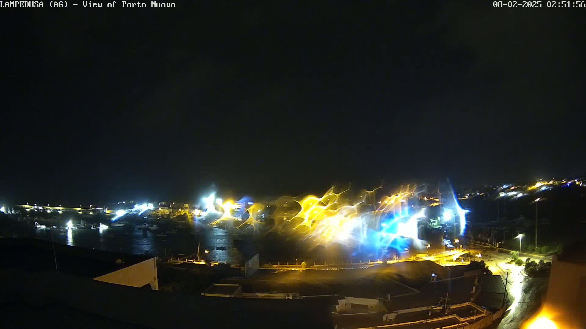 A nighttime view of a harbor with boats, illuminated buildings, and streaks of light across the water under a dark sky.