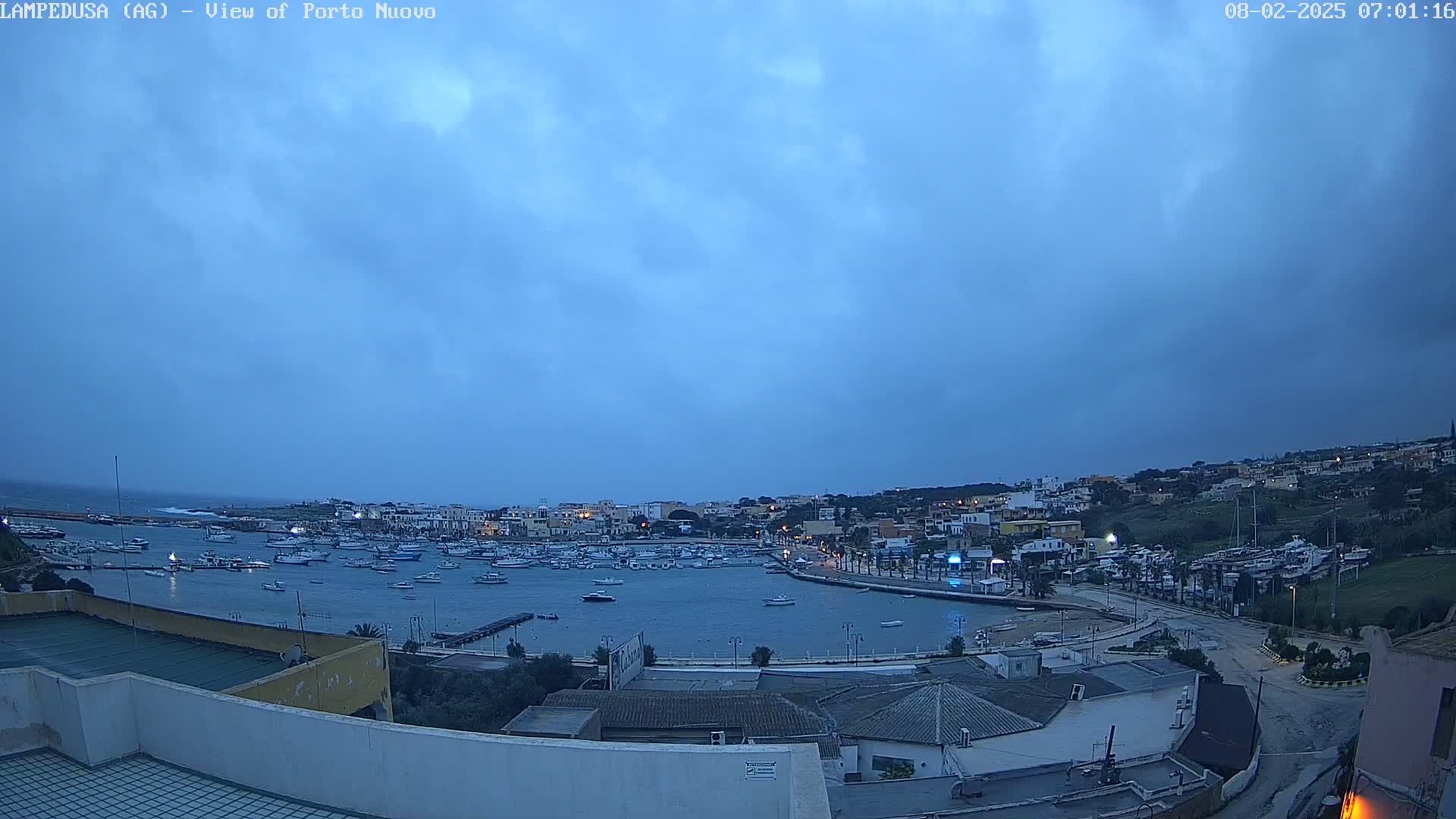 An overcast, pre-dawn view of a harbor filled with boats, next to a town built on a hillside.
