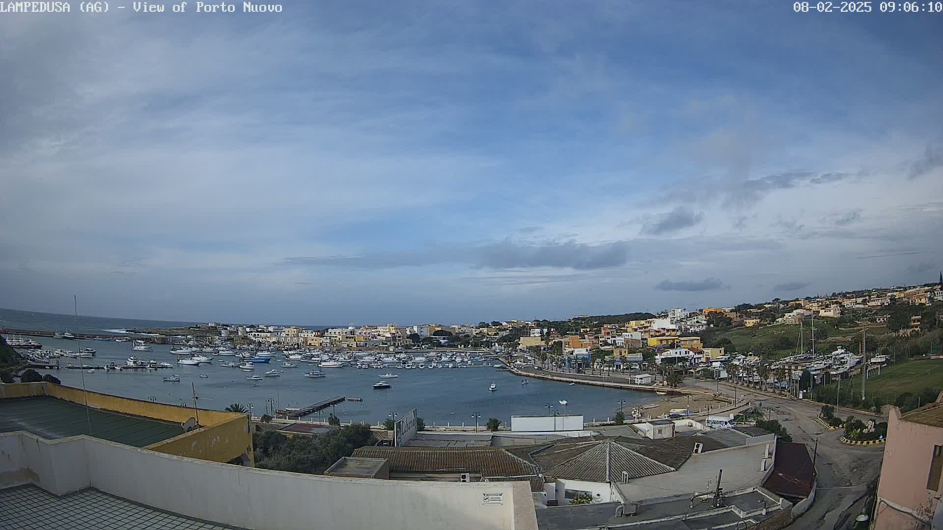A harbor filled with numerous boats is nestled within a town of low-rise buildings under a mostly clear sky with some scattered clouds.