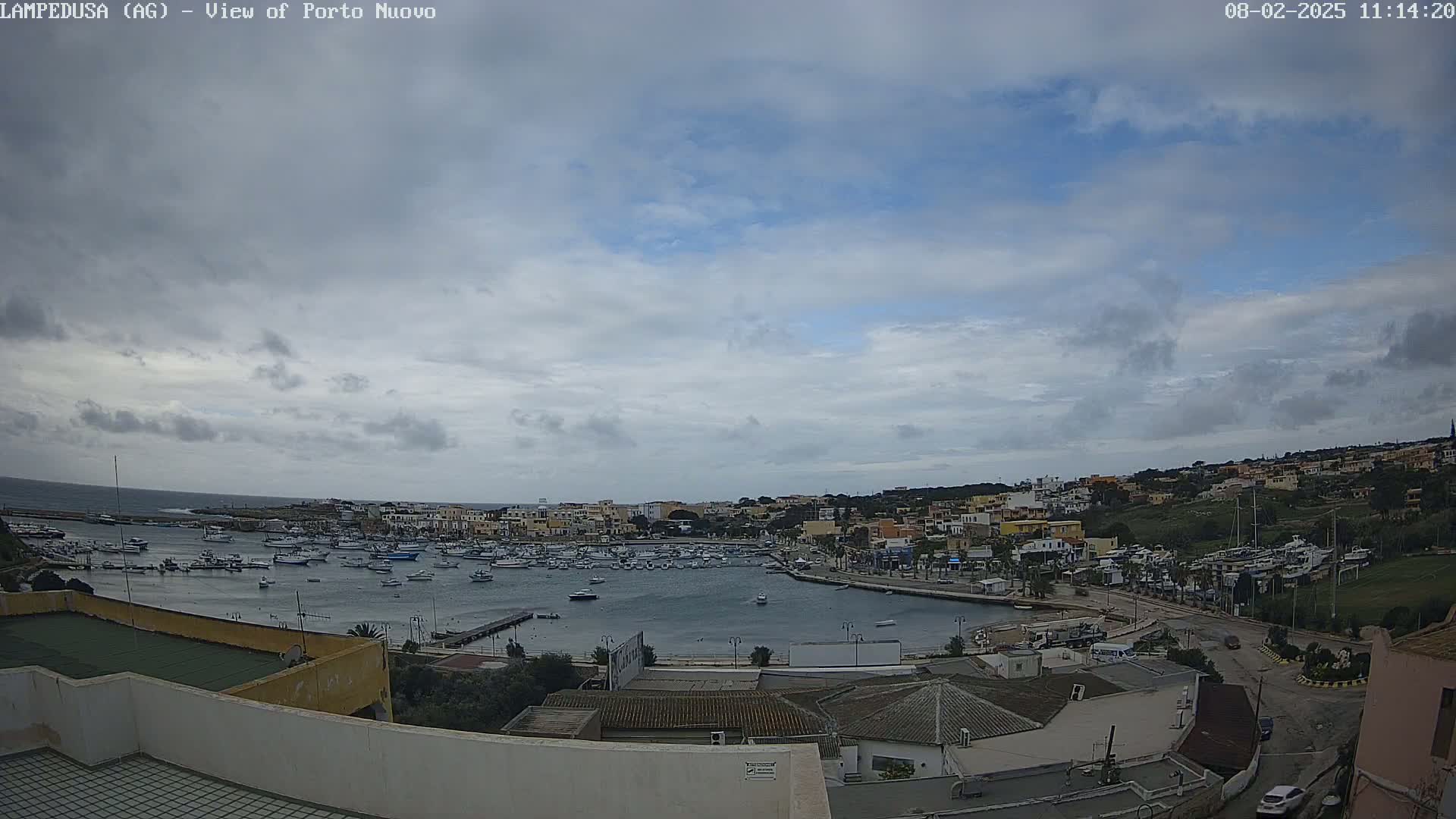 A mostly cloudy sky overlooks a harbor filled with numerous boats and a town built on a hillside.