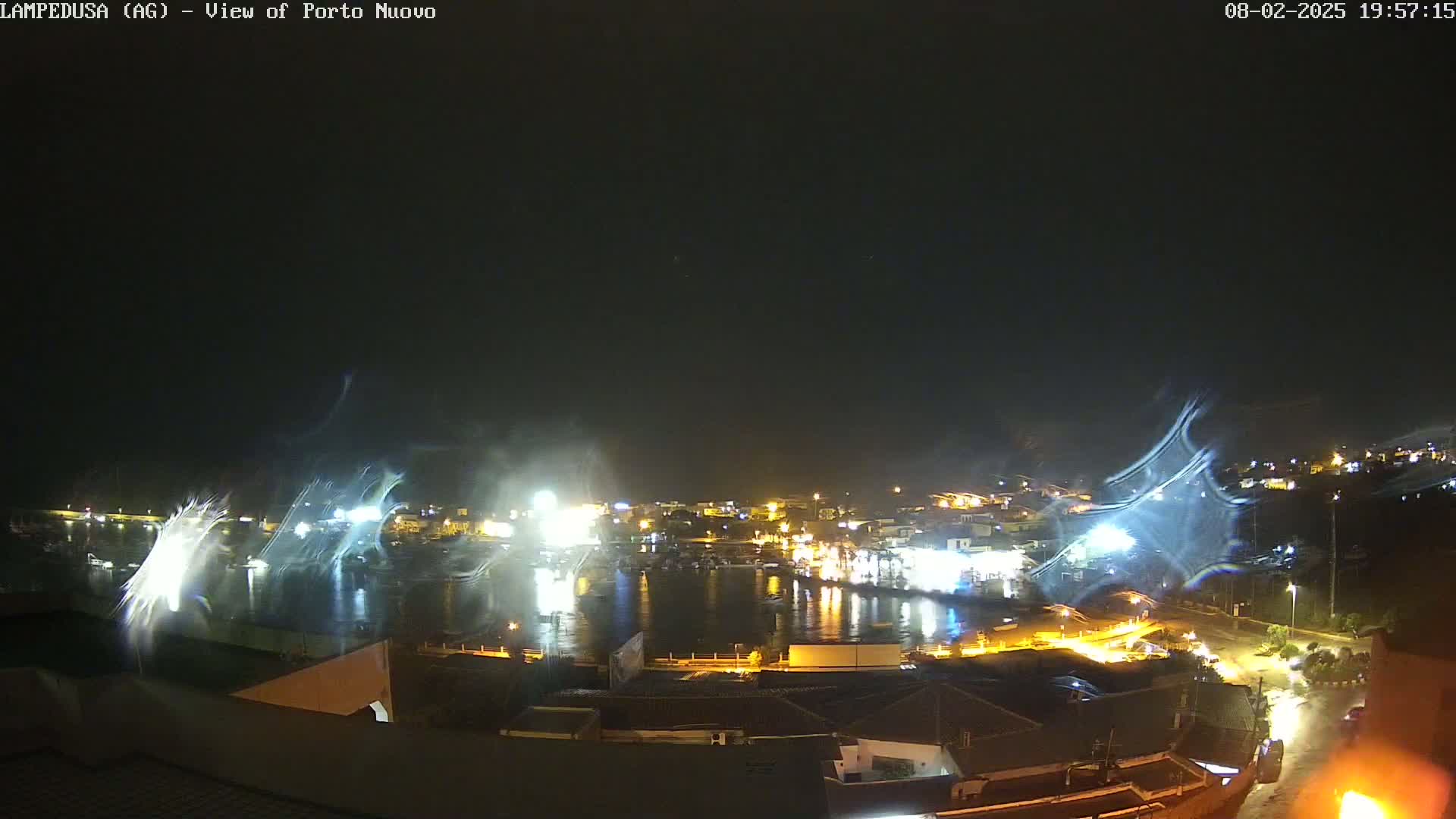 A nighttime view of a harbor town with its lights illuminated, under a dark sky with streaks of what appear to be windblown spray or mist.