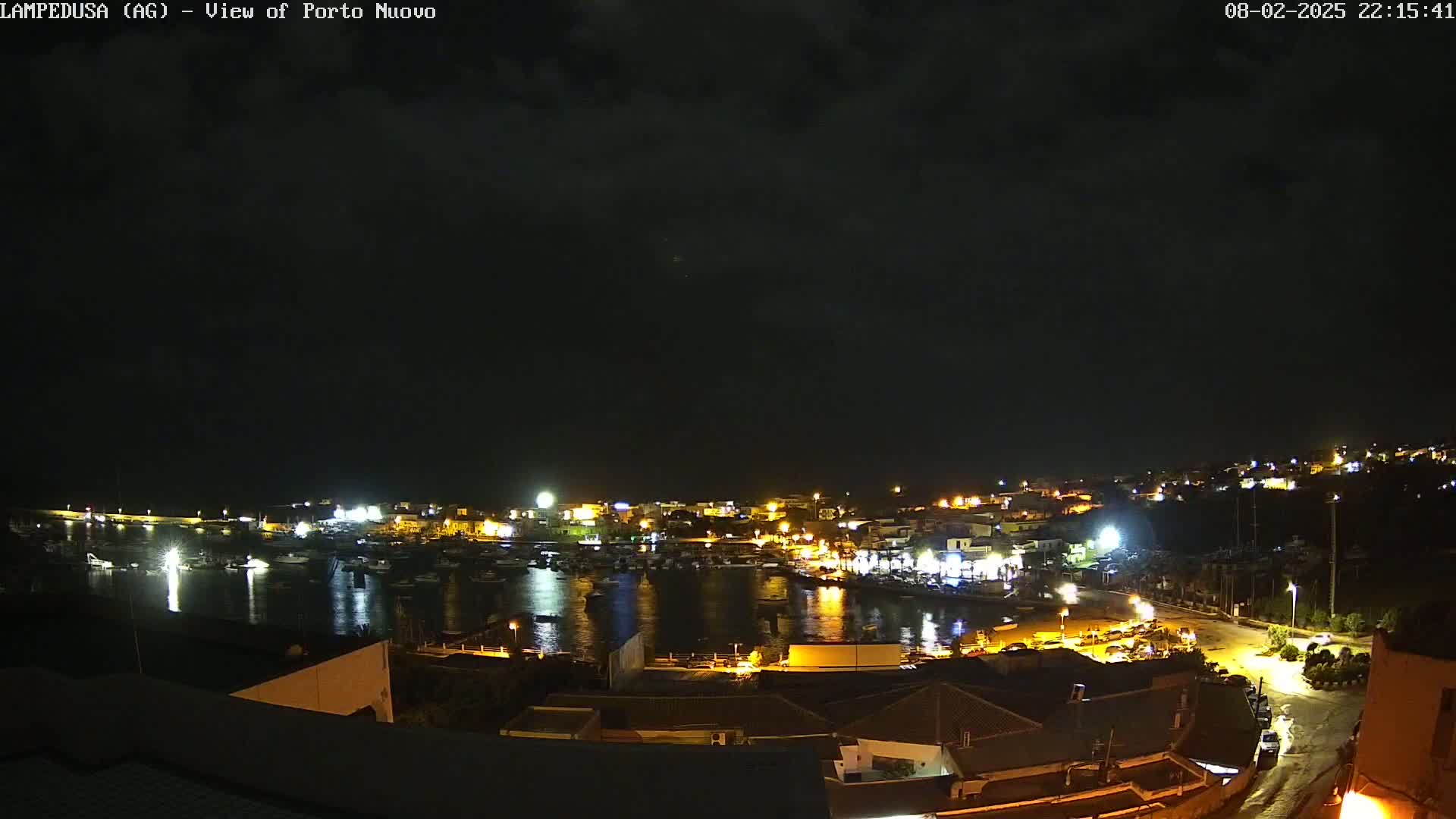 A nighttime view of a harbor filled with boats, surrounded by a town with illuminated buildings under a dark, cloudy sky.