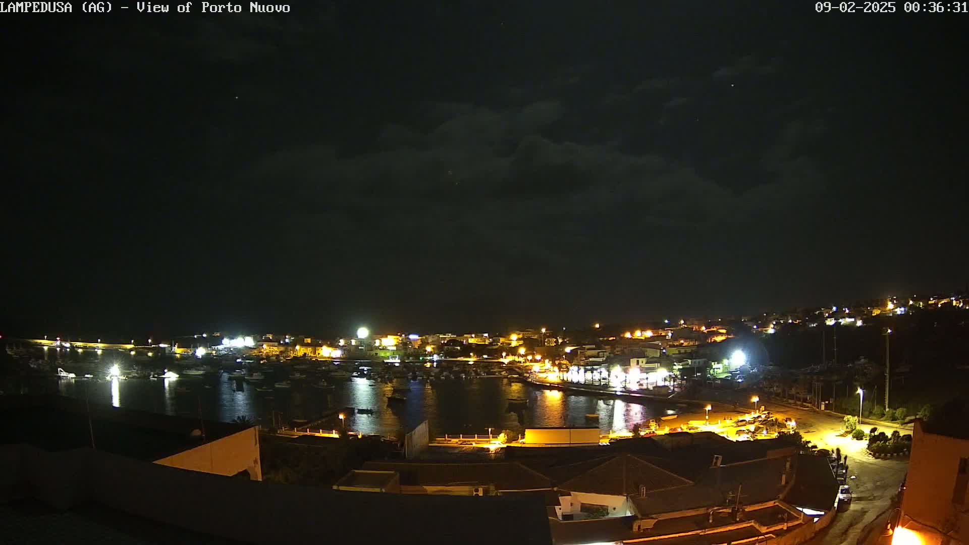 A nighttime view of a harbor town with many lights illuminated under a partly cloudy sky.