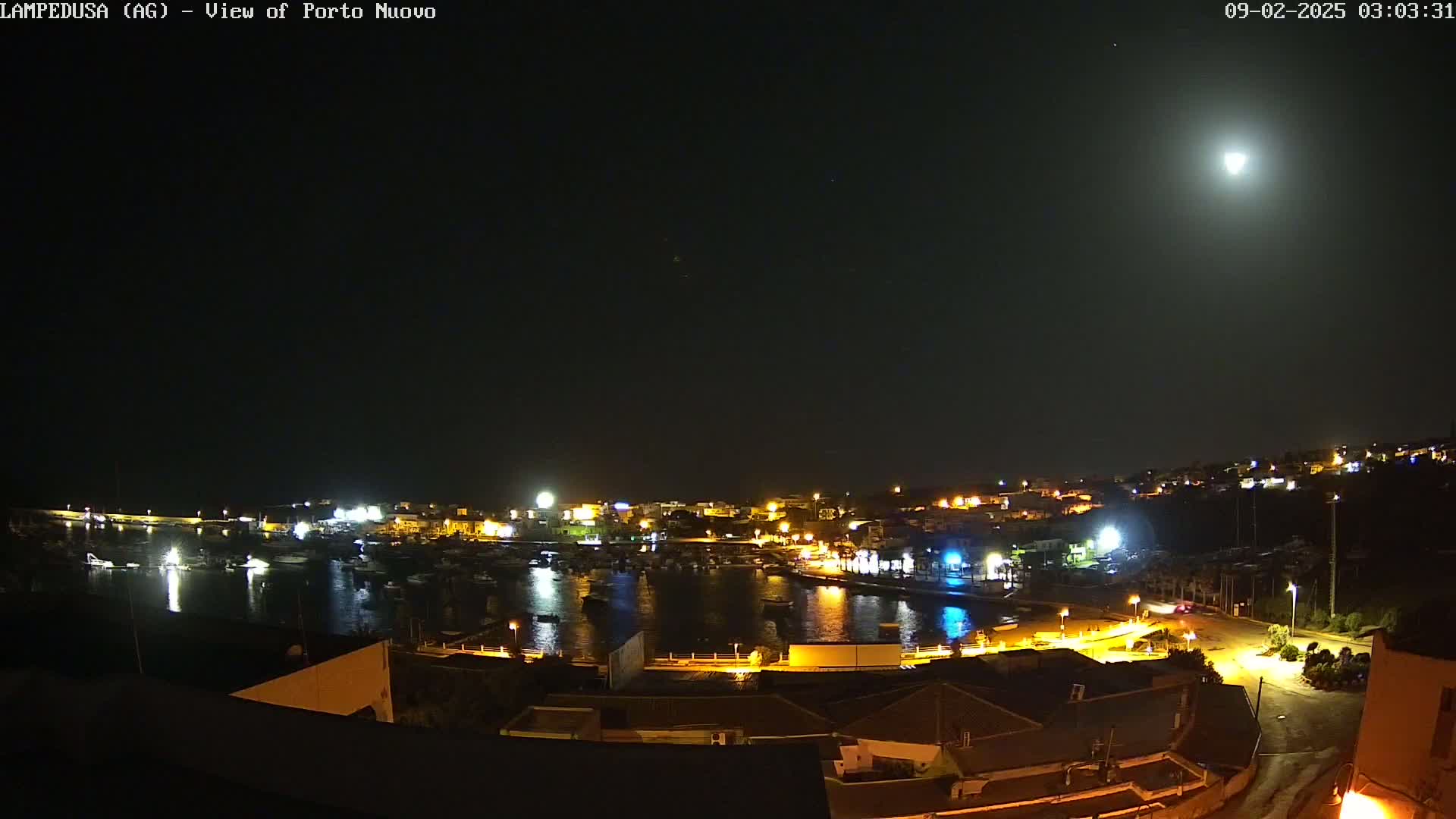 A brightly lit harbor town is nestled on a hillside under a clear night sky illuminated by a full moon.