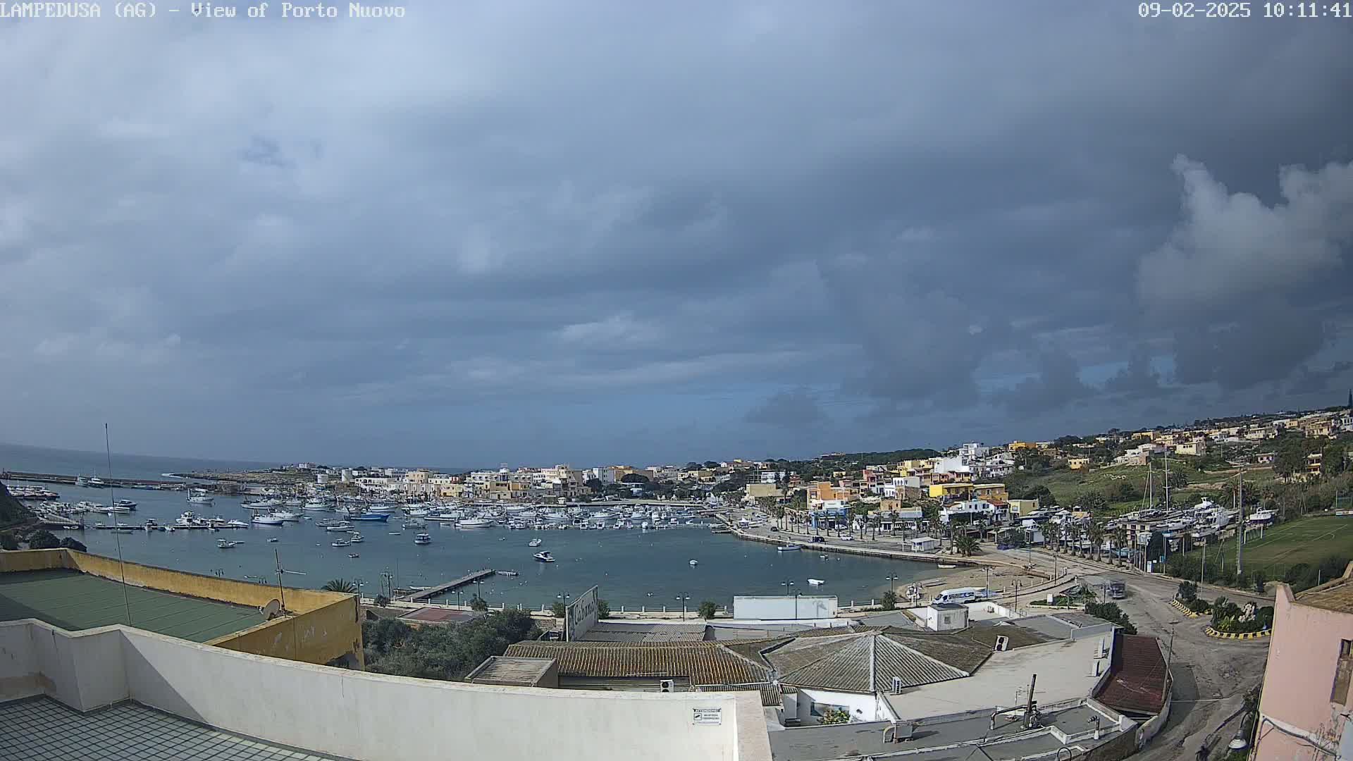 An overcast sky hangs over a harbor filled with numerous boats, surrounded by a town of low-rise buildings nestled against a hillside.