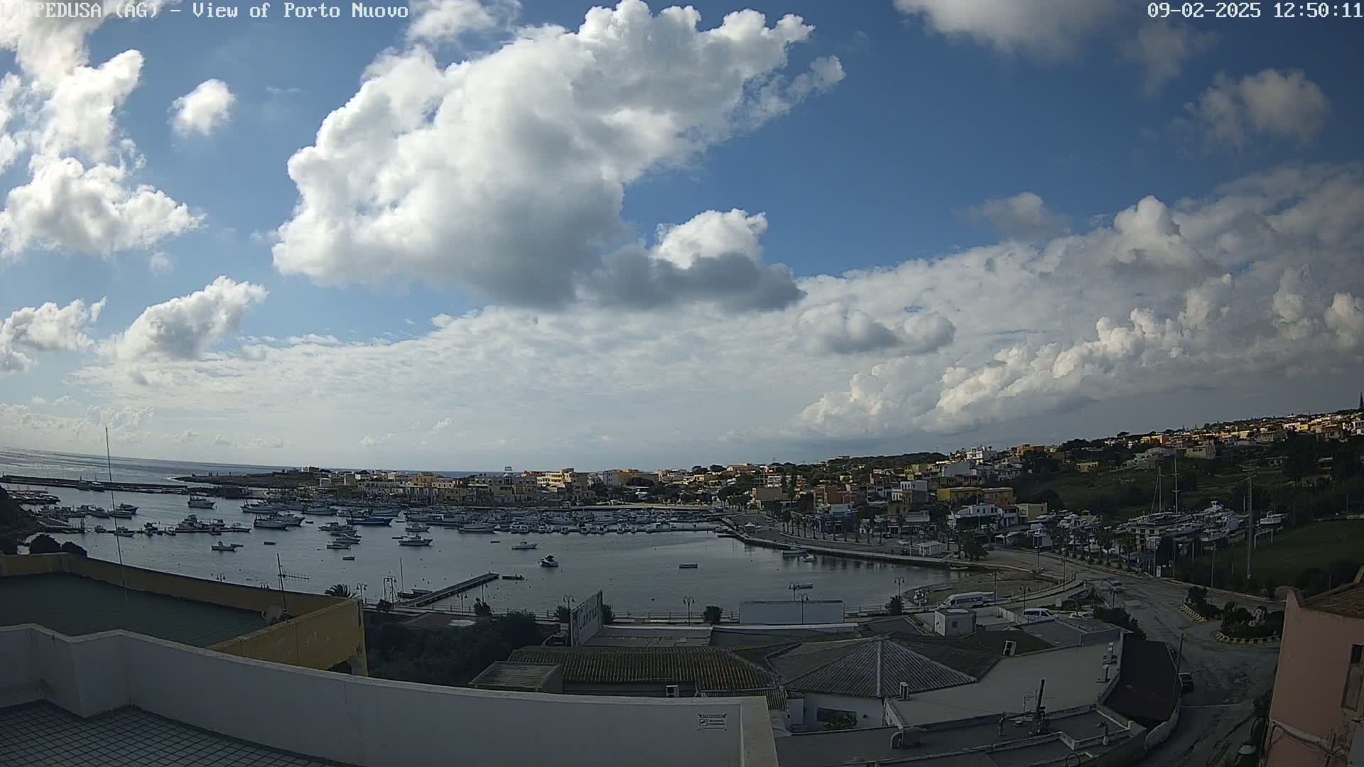 A partly cloudy sky overlooks a harbor filled with numerous boats and a town nestled against a hillside.