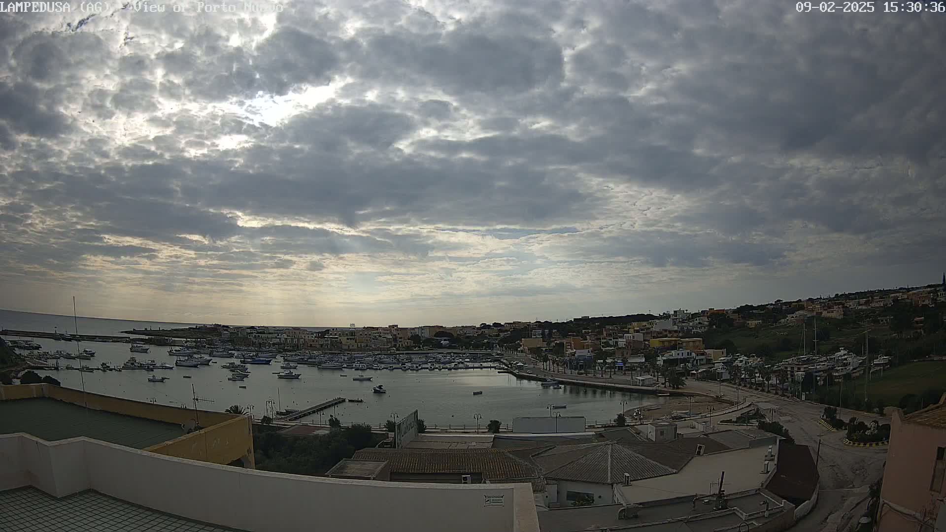 An overcast sky hangs over a harbor filled with boats, and a town of low-rise buildings nestled against a hillside.