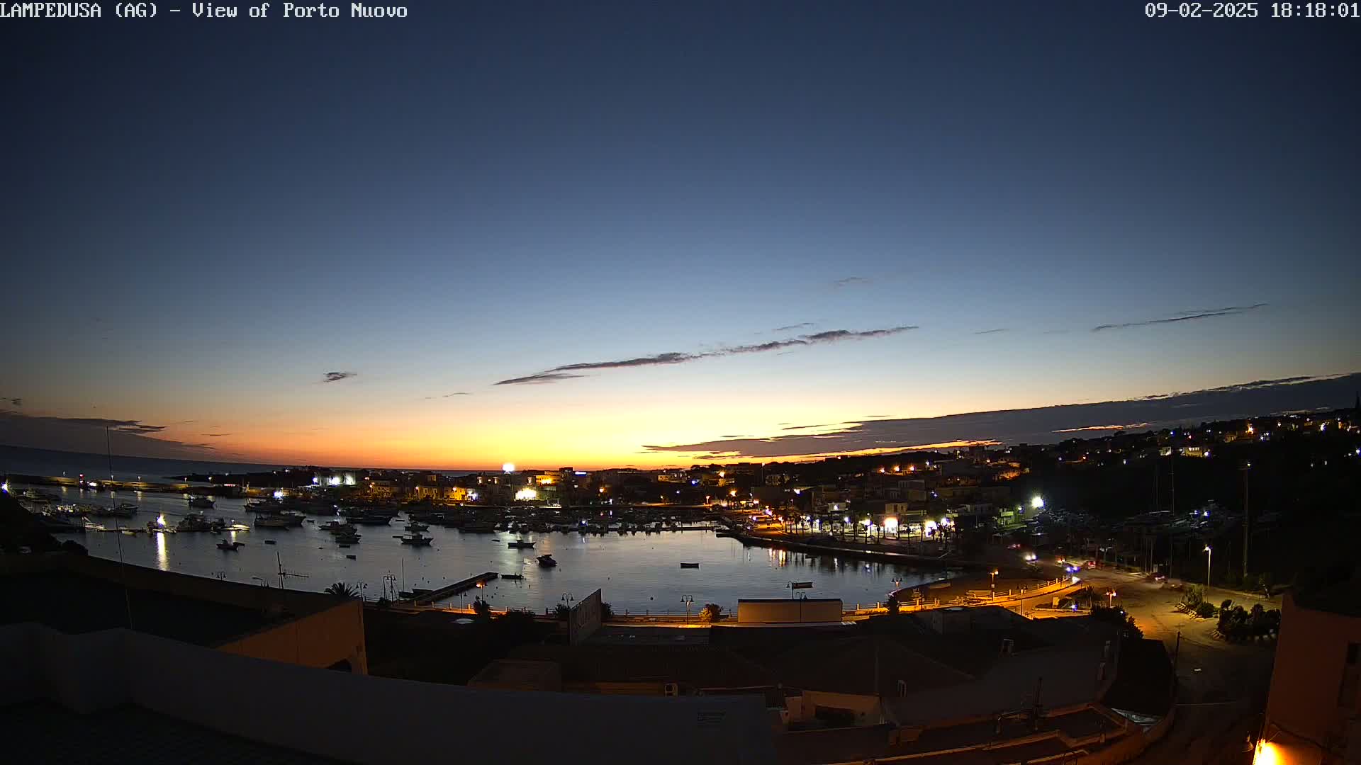 A nighttime harbor scene shows a calm body of water filled with boats, surrounded by a town with illuminated buildings, all under a twilight sky with hints of orange from the setting sun.