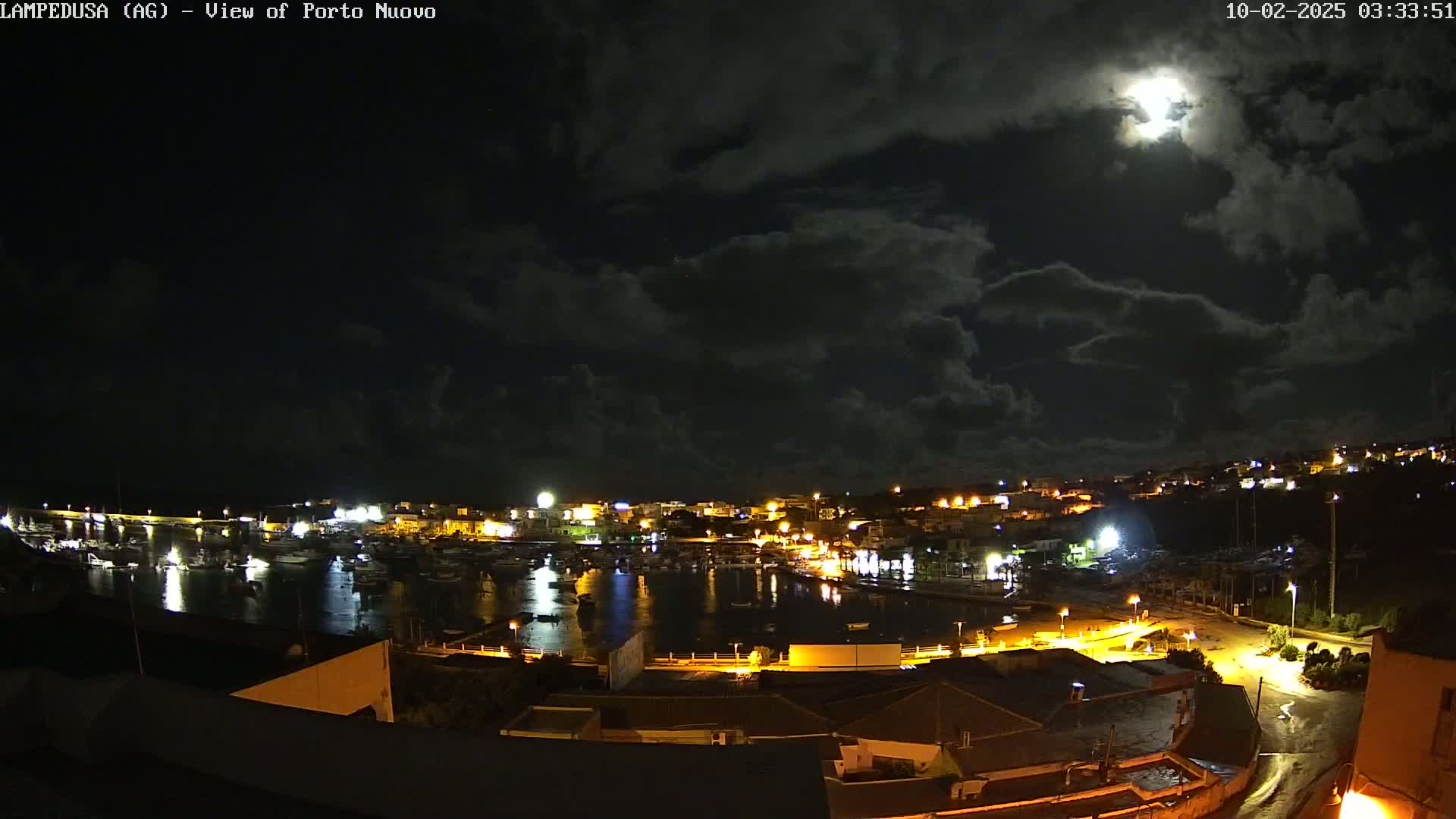 A nighttime view of a harbor town illuminated by lights under a partly cloudy sky with a bright moon.