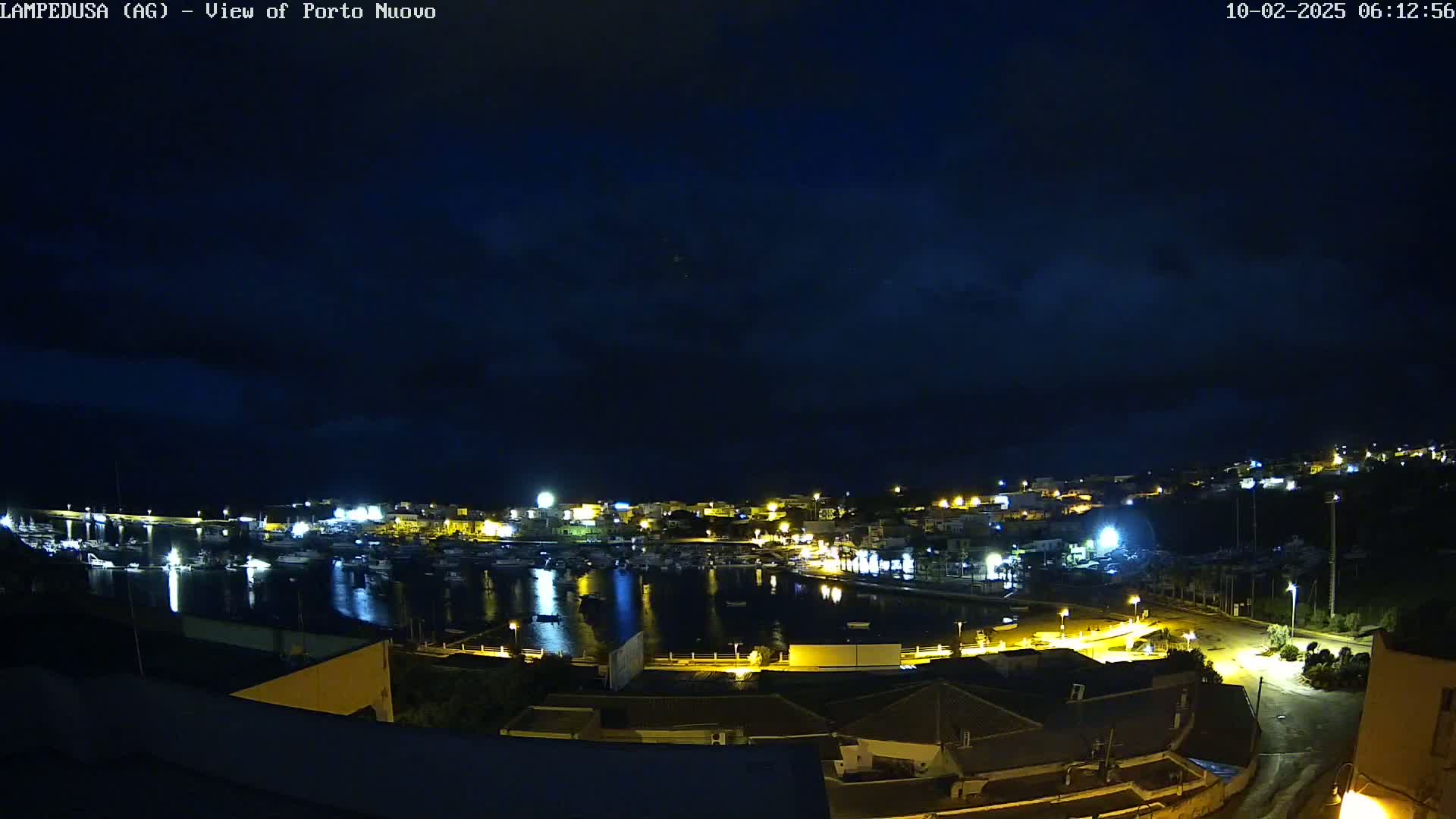 A nighttime, slightly cloudy view of a harbor town with boats in the water and illuminated buildings along the shoreline.