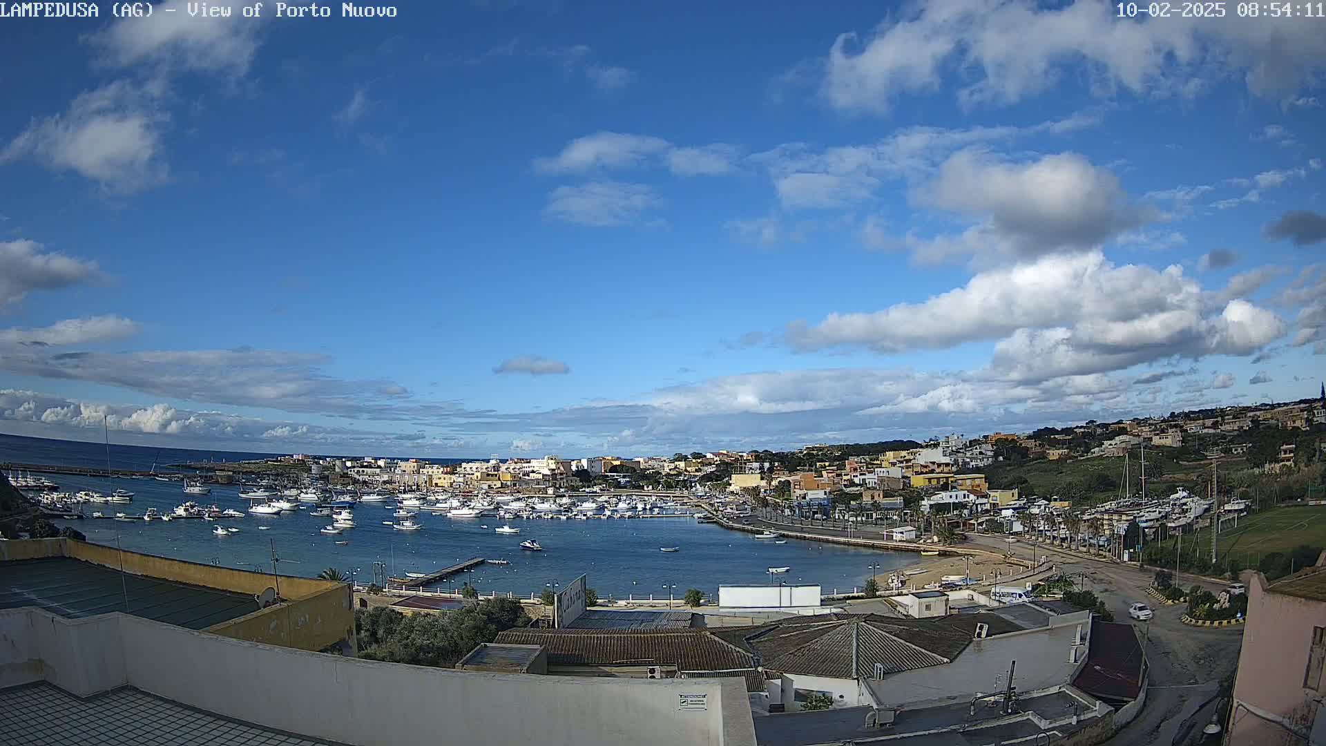 A harbor filled with boats is nestled in a town on a coast under a partly cloudy sky.