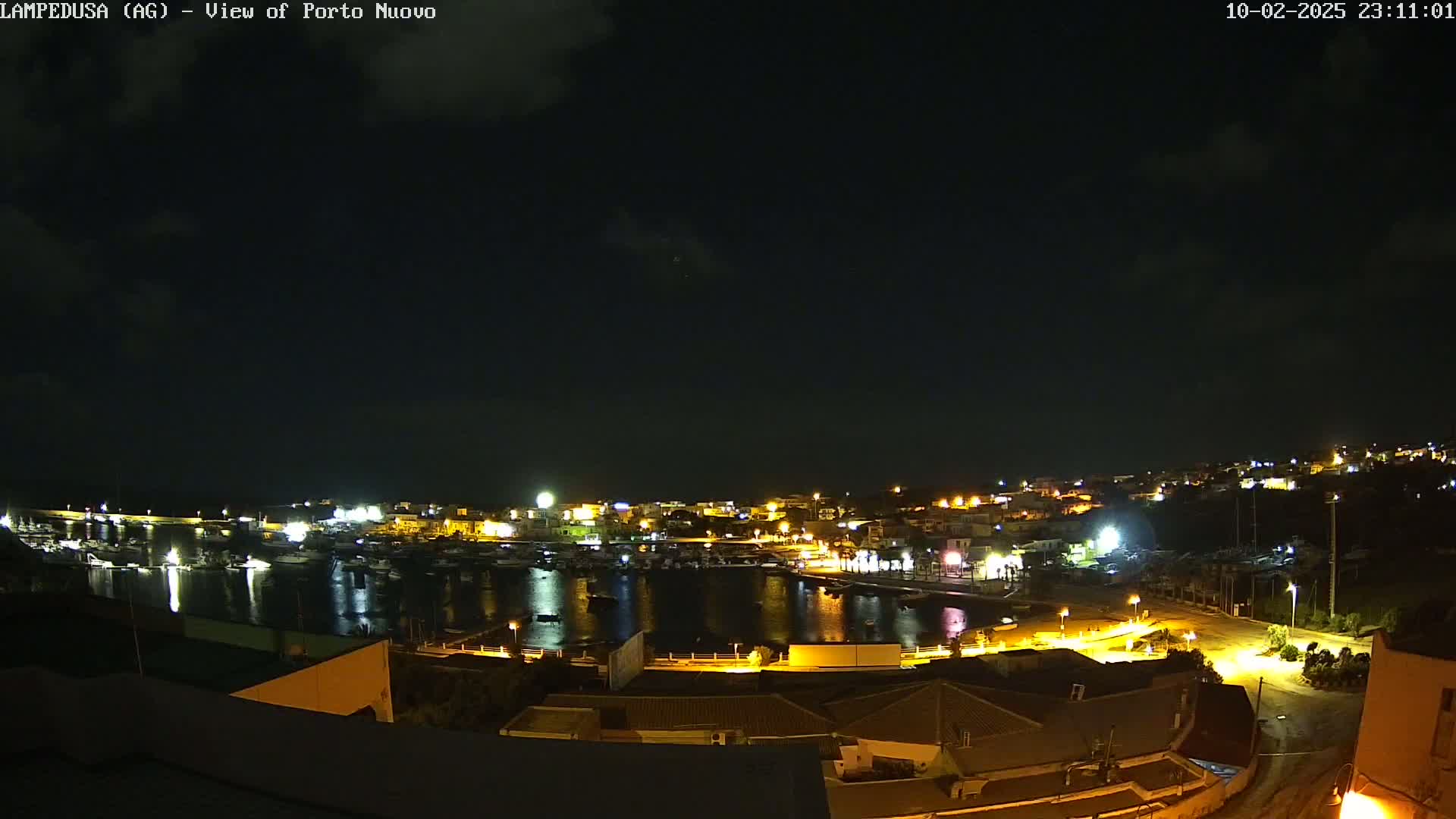 A nighttime view of a harbor town with boats in the water and buildings lit up under a partly cloudy sky.