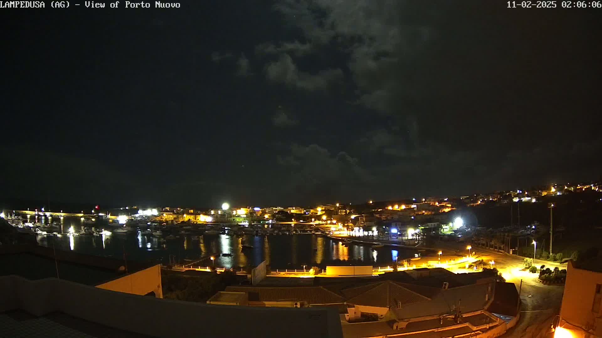 A nighttime view of a harbor town with boats in the water under a partly cloudy sky.