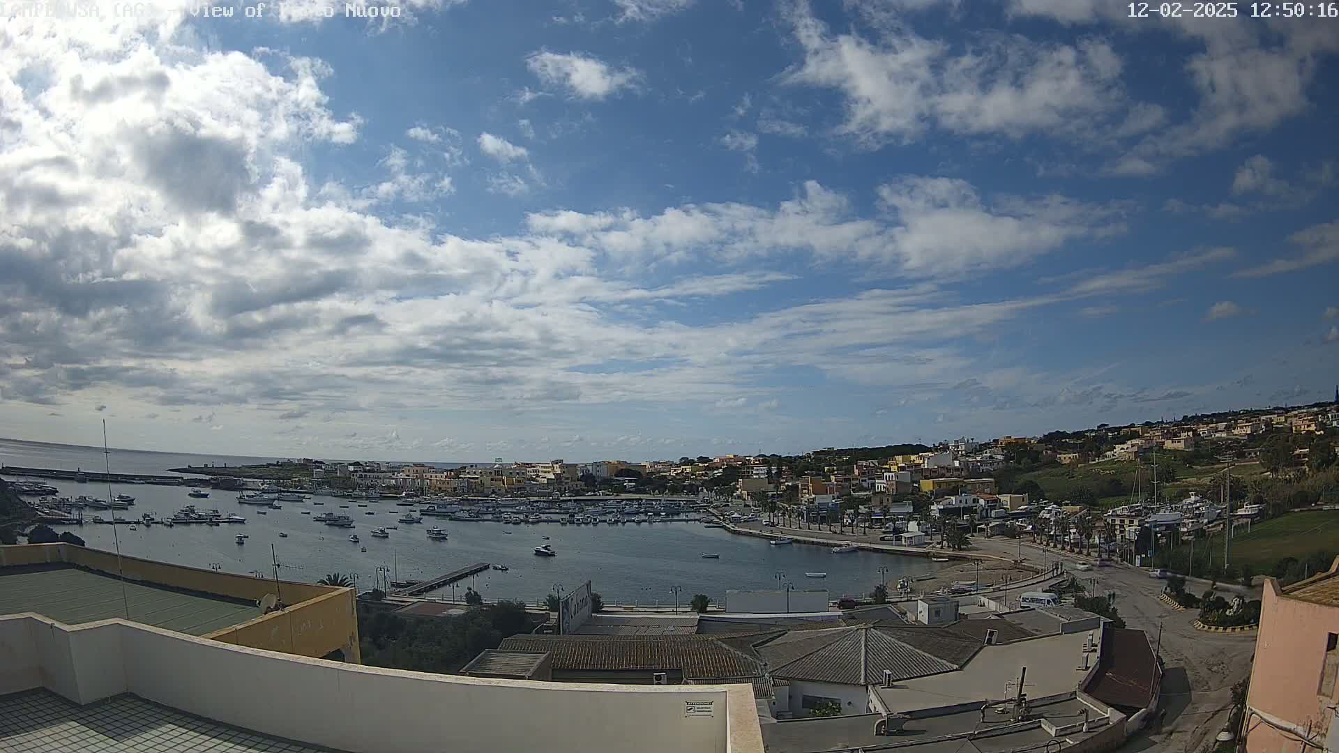A partly cloudy sky overlooks a harbor filled with boats, adjacent to a town built on a hillside.
