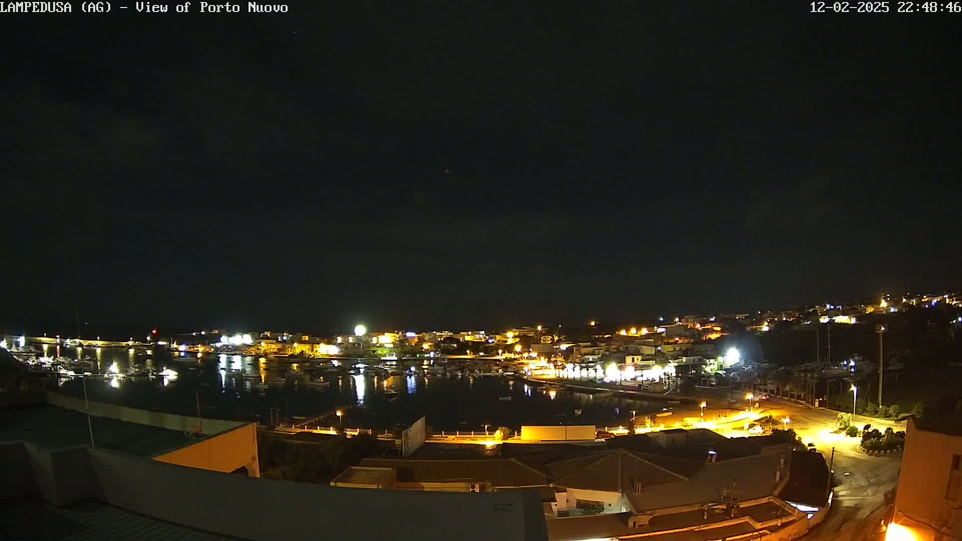 A nighttime view of a harbor filled with boats, surrounded by illuminated buildings and streets under a dark, clear sky.
