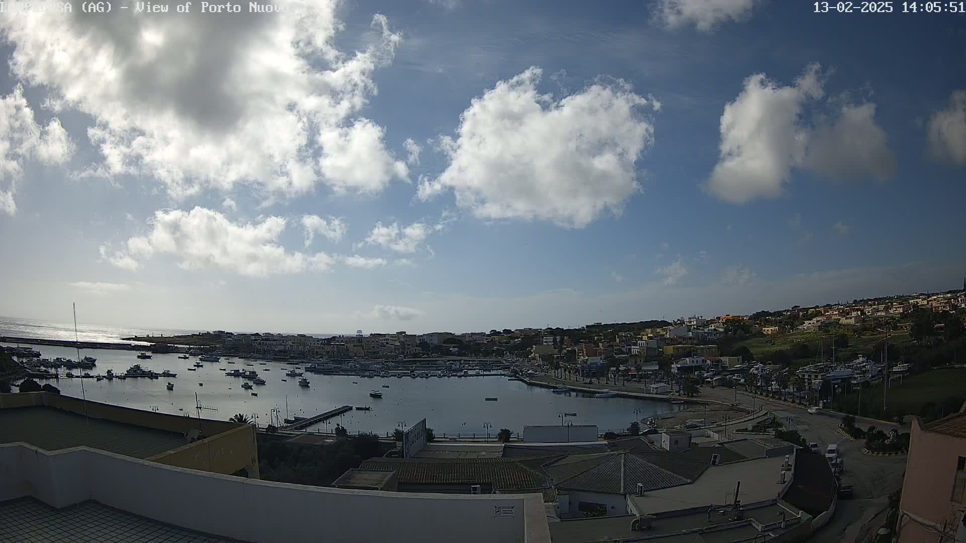 A partly cloudy sky overlooks a harbor filled with boats and a town with buildings extending along the coast.