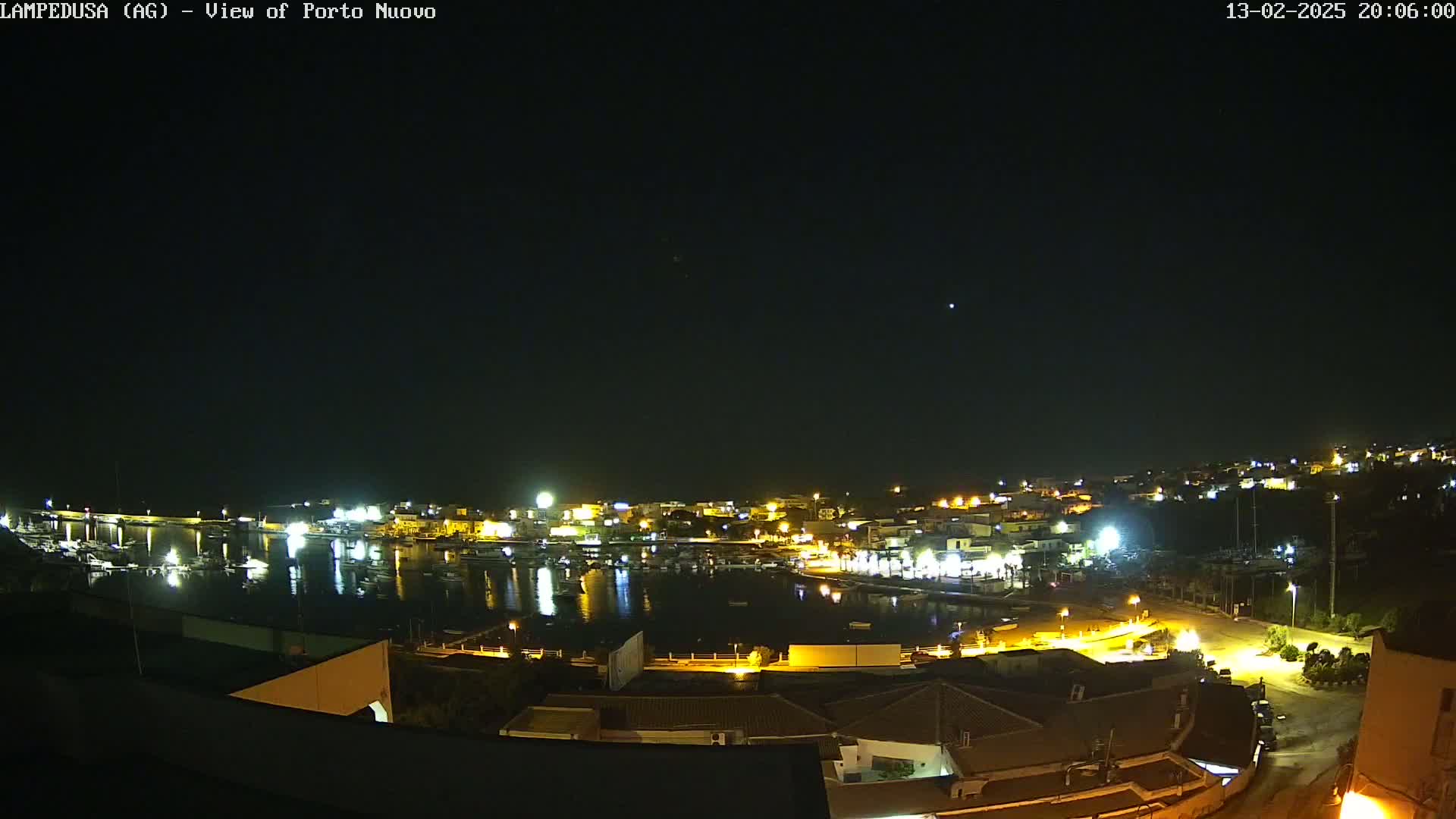 A nighttime, clear-sky view of a harbor town with brightly lit buildings and boats, seen from an elevated vantage point.