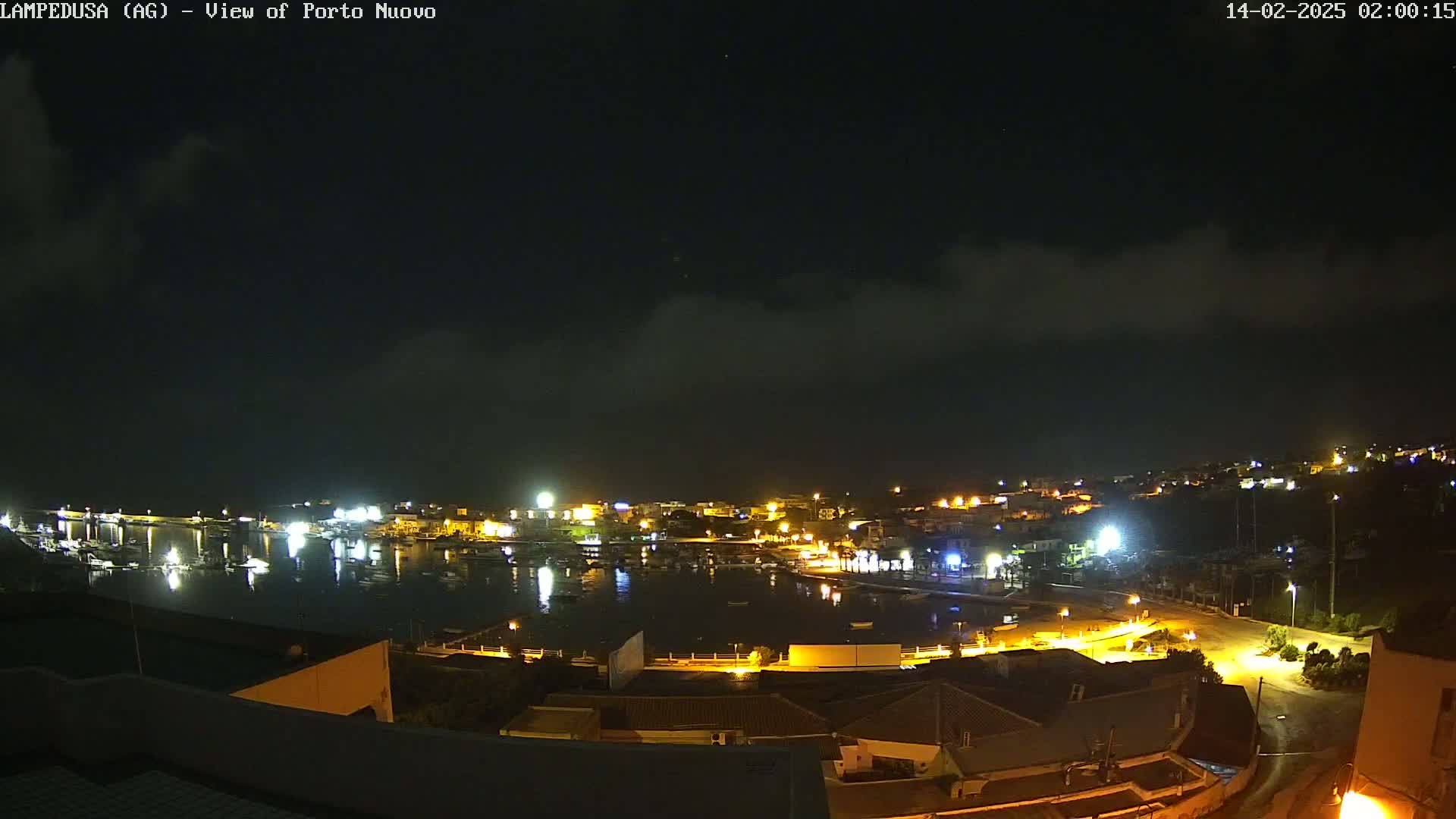 A nighttime view of a harbor town with many lights illuminating the waterfront and surrounding buildings under a mostly cloudy sky.
