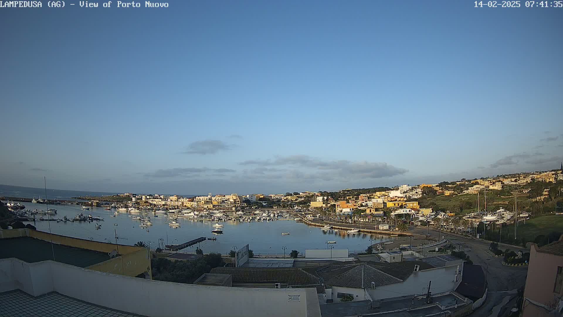 A harbor filled with boats is surrounded by a town of low-lying buildings under a mostly clear, light-blue sky.