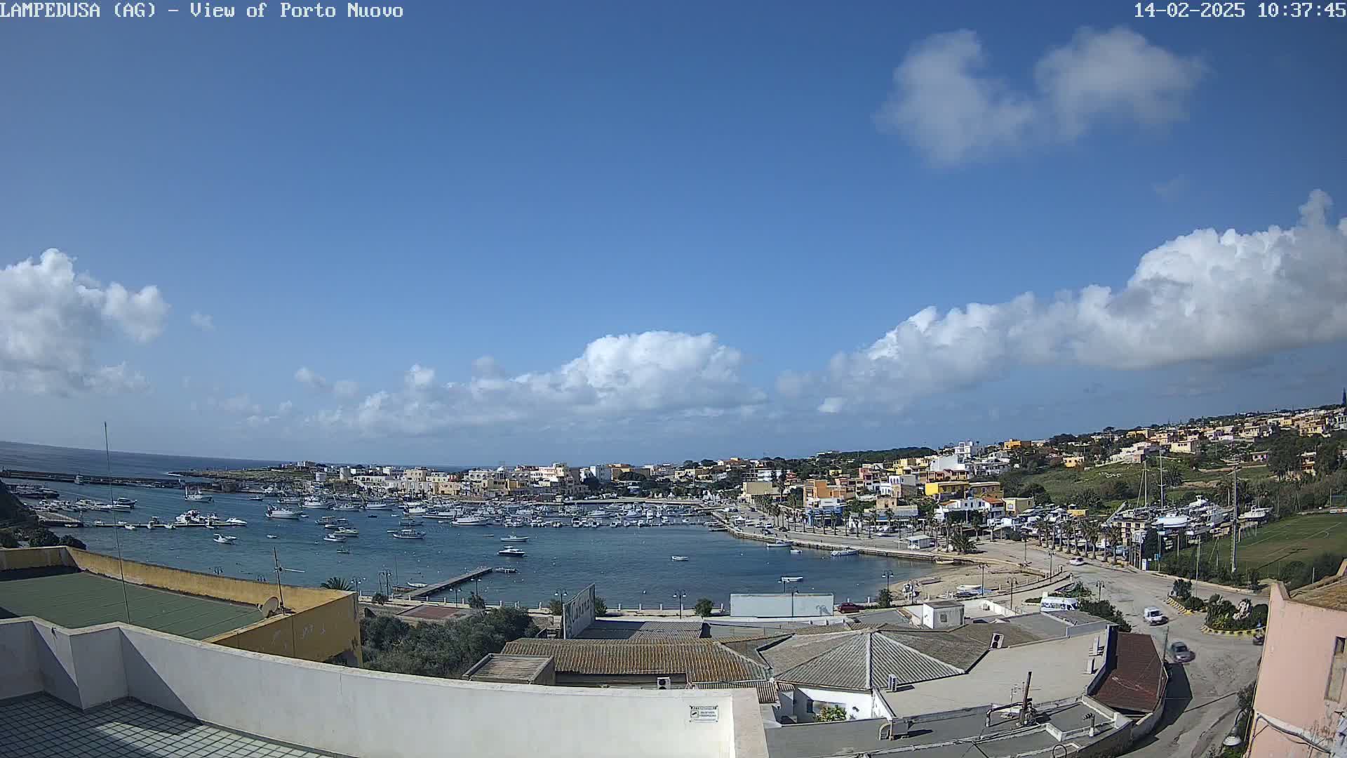 A mostly sunny day shows a harbor filled with boats, next to a town of low-rise buildings, under a partly cloudy sky.