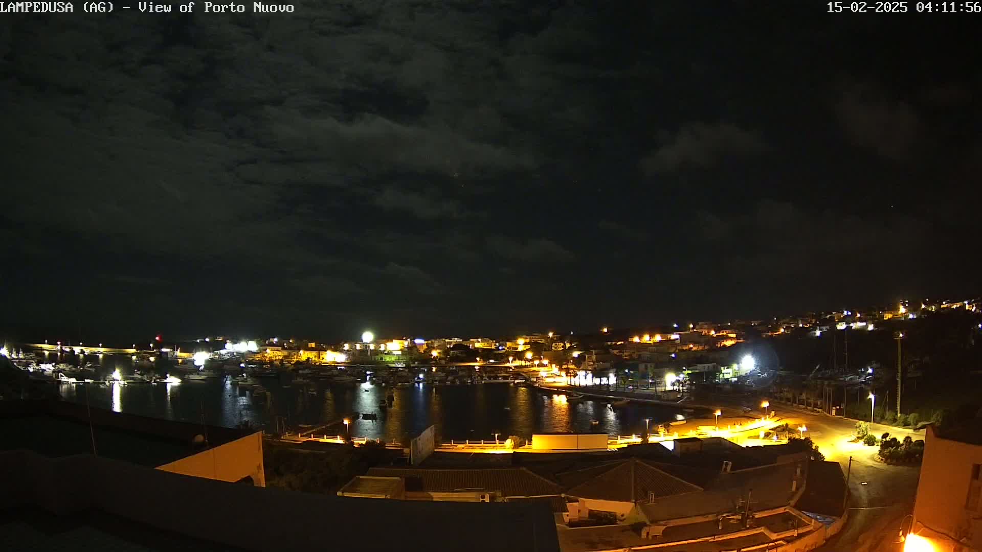 A nighttime view of a harbor town, illuminated by lights, under a partly cloudy sky.