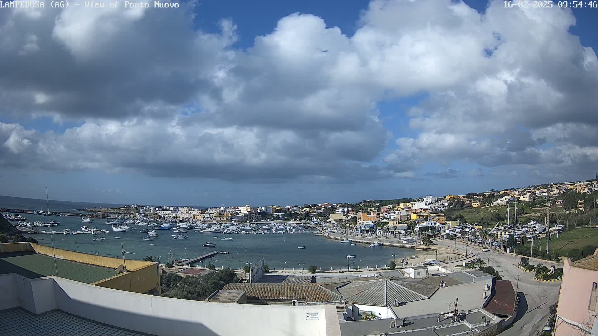 A harbor filled with boats is surrounded by a town situated on a hill under a partly cloudy sky.