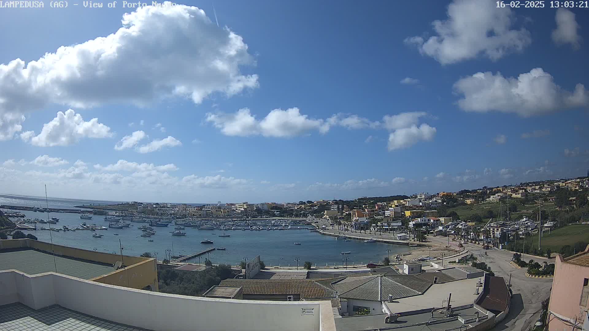 A partly cloudy blue sky overlooks a harbor filled with boats and a town of low-rise buildings on a hillside.