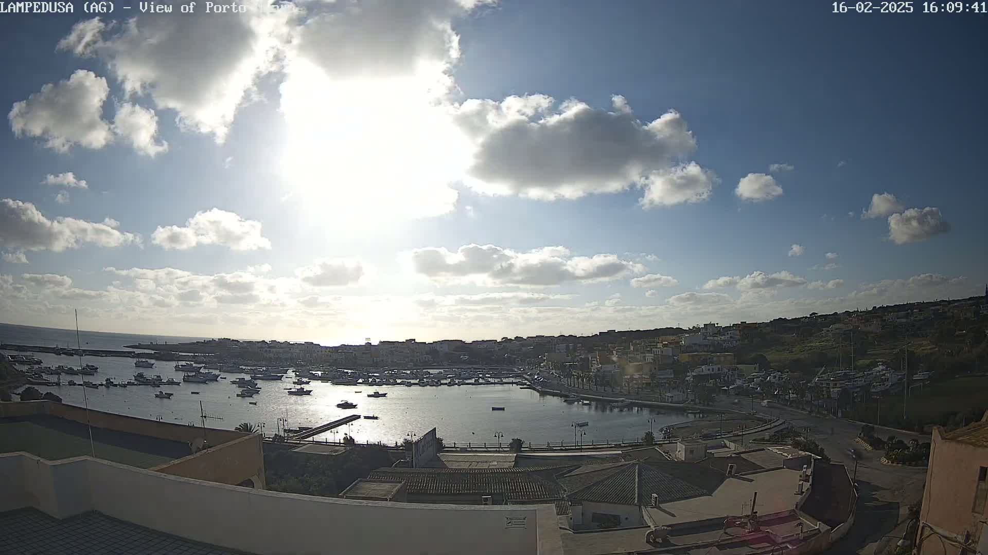 A partly sunny day overlooks a harbor filled with boats, surrounded by buildings and hills.