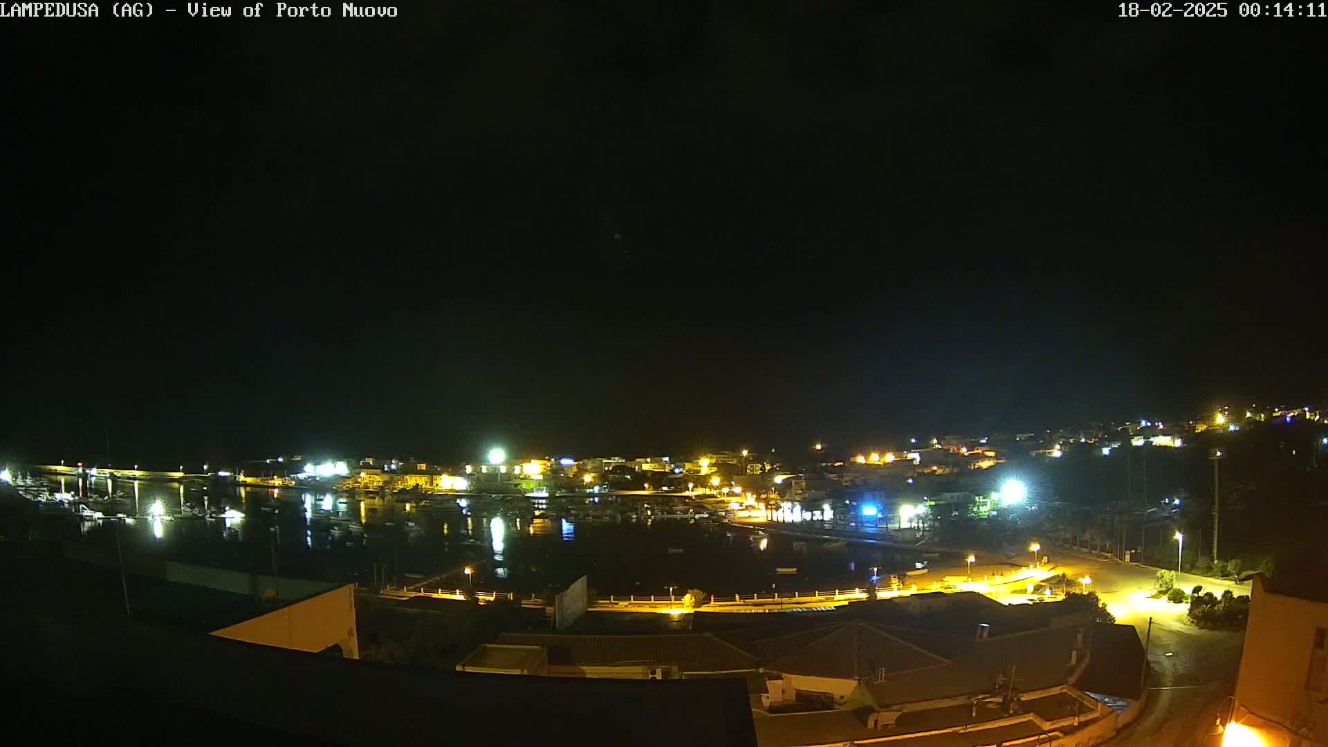 A nighttime, aerial view shows a harbor town with its lights illuminating the water and surrounding buildings under a clear, dark sky.
