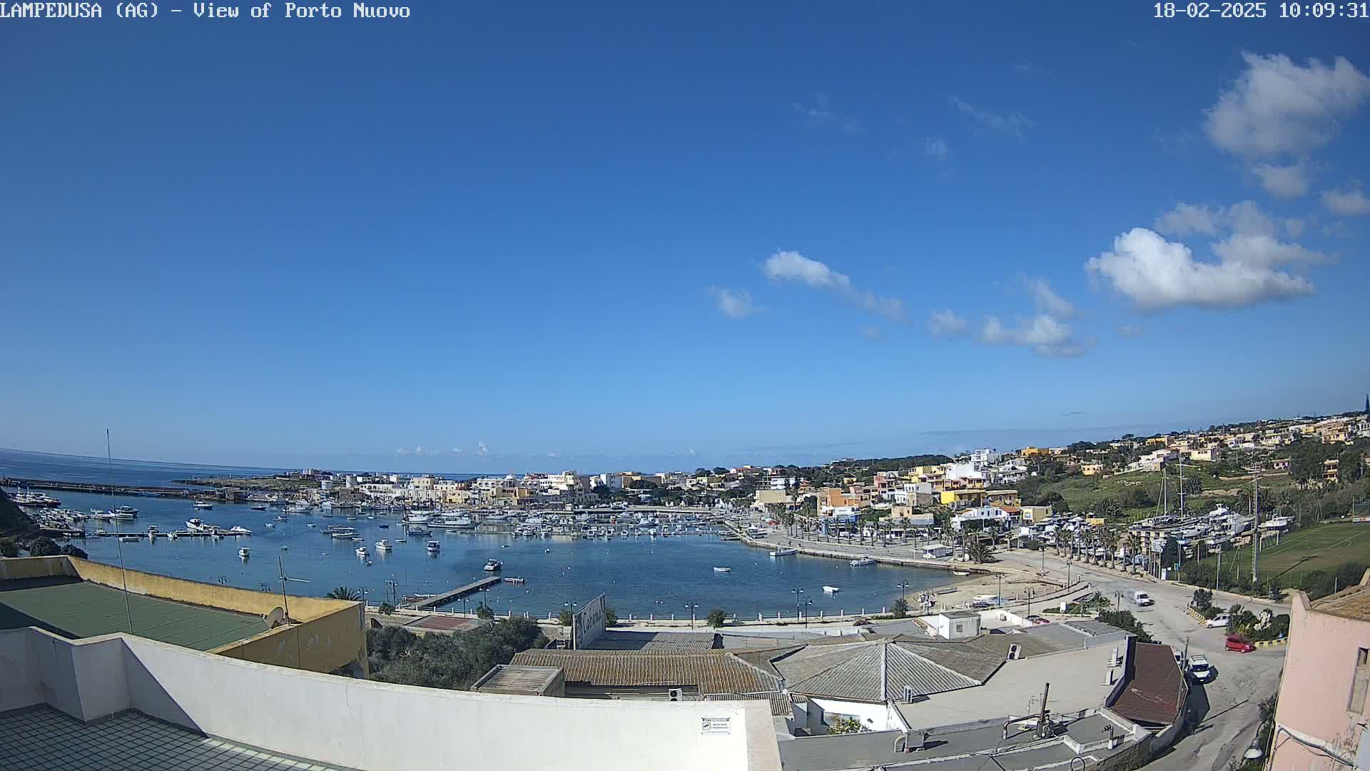 A mostly sunny day shows a harbor filled with boats, surrounded by a town of low buildings under a mostly clear blue sky.