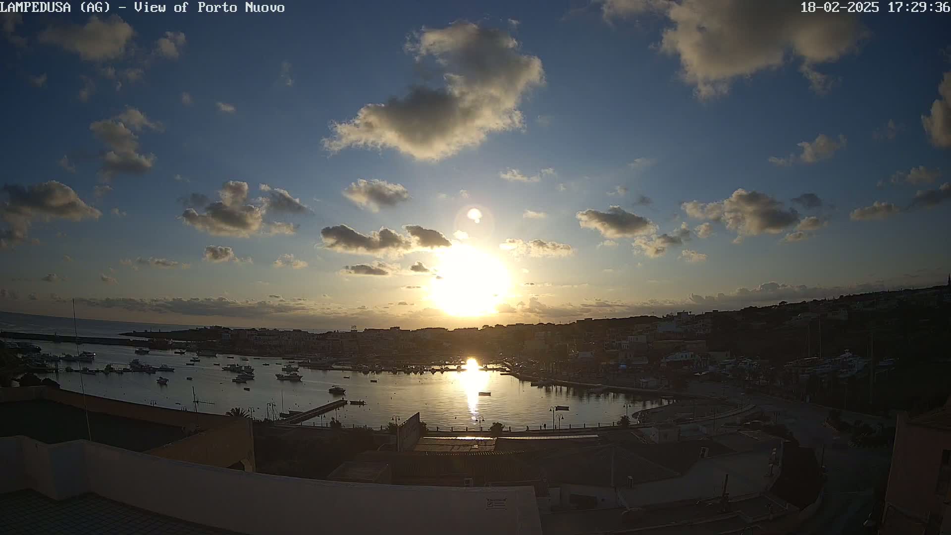 A partly cloudy sunset over a harbor filled with boats and a town nestled on a hillside.