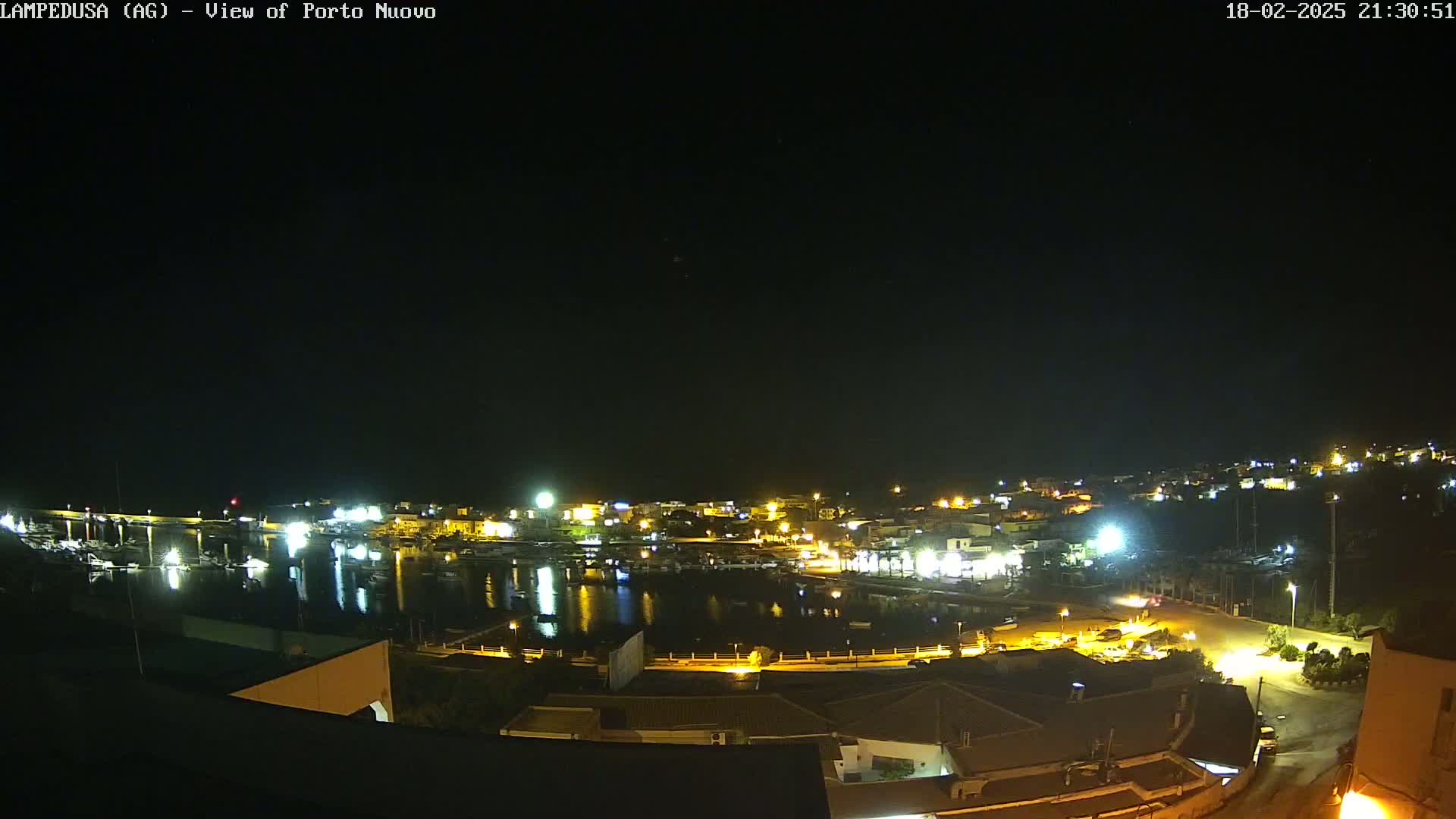 A nighttime view of a harbor town, with many lights illuminating buildings and boats along the water under a clear, dark sky.