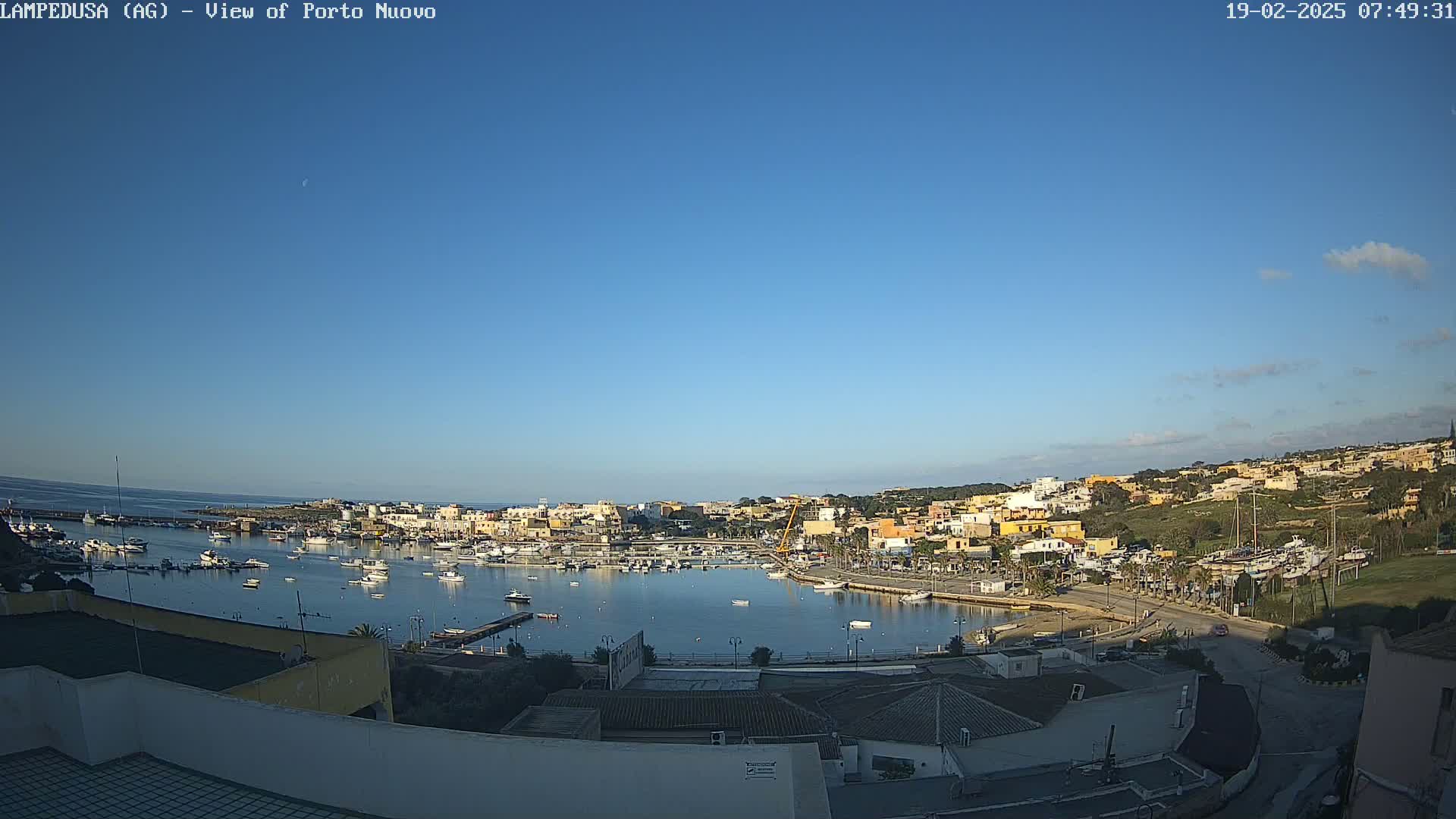 A clear, sunny day overlooks a harbor filled with boats, nestled beside a town of low-rise buildings against a backdrop of hills.