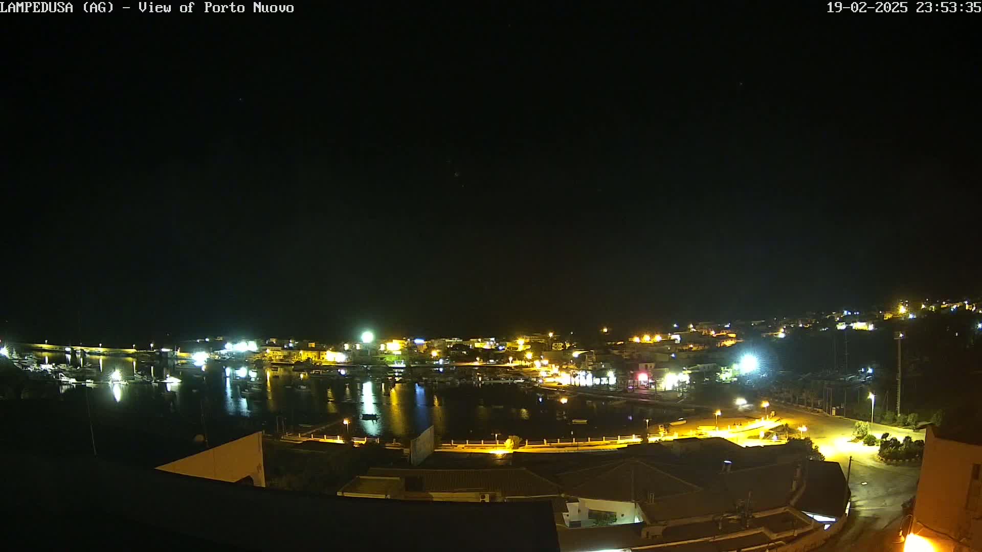 A nighttime, high-angle view of a harbor town with many lights illuminating the buildings and the water, under a clear, dark sky.
