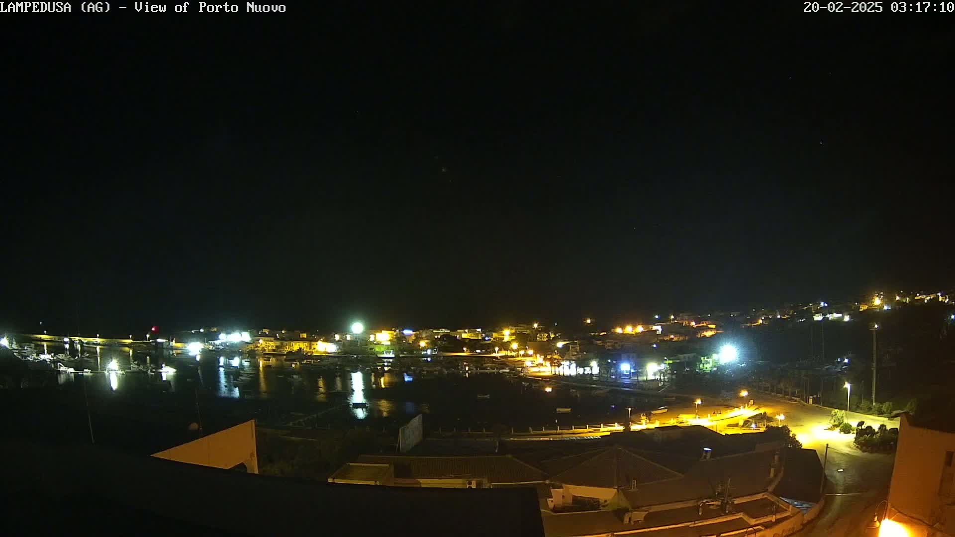A nighttime view of a harbor town with many lights illuminating buildings and boats under a clear, dark sky.