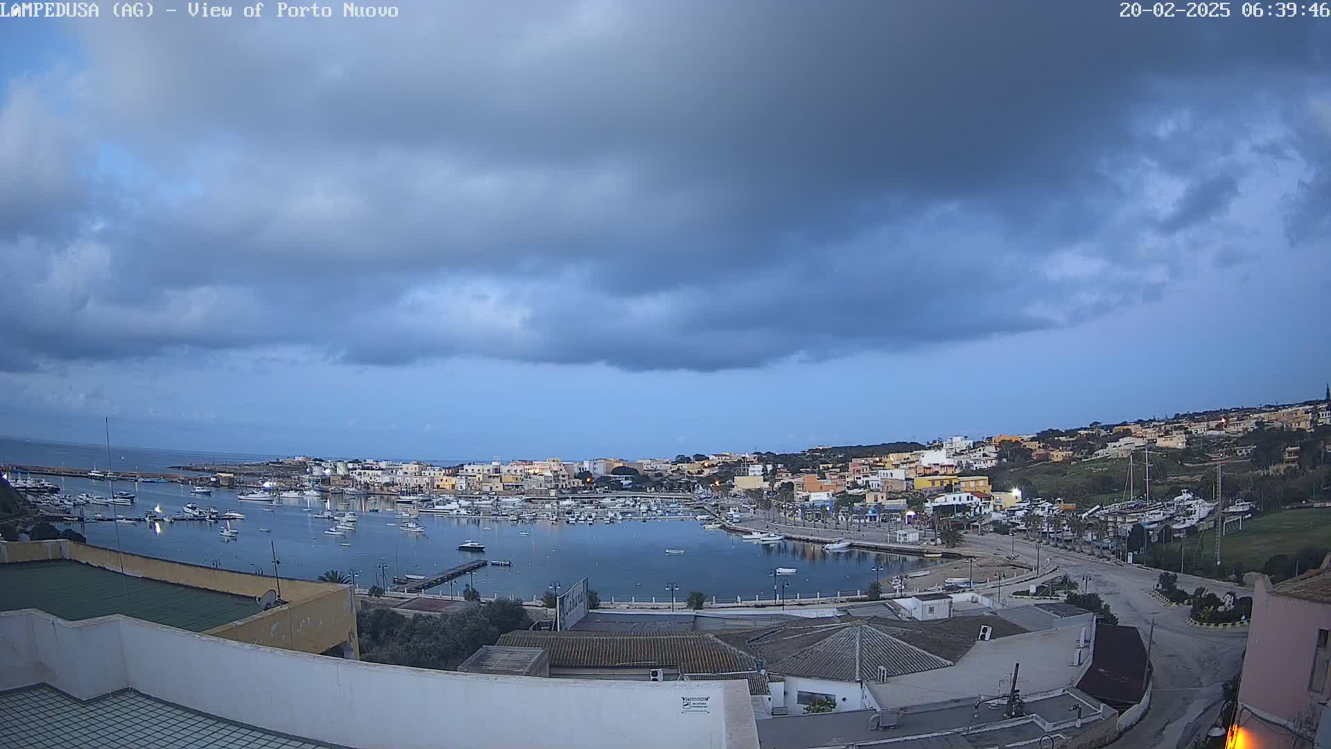 A harbor filled with boats is nestled in a town on a hillside under a mostly cloudy sky.