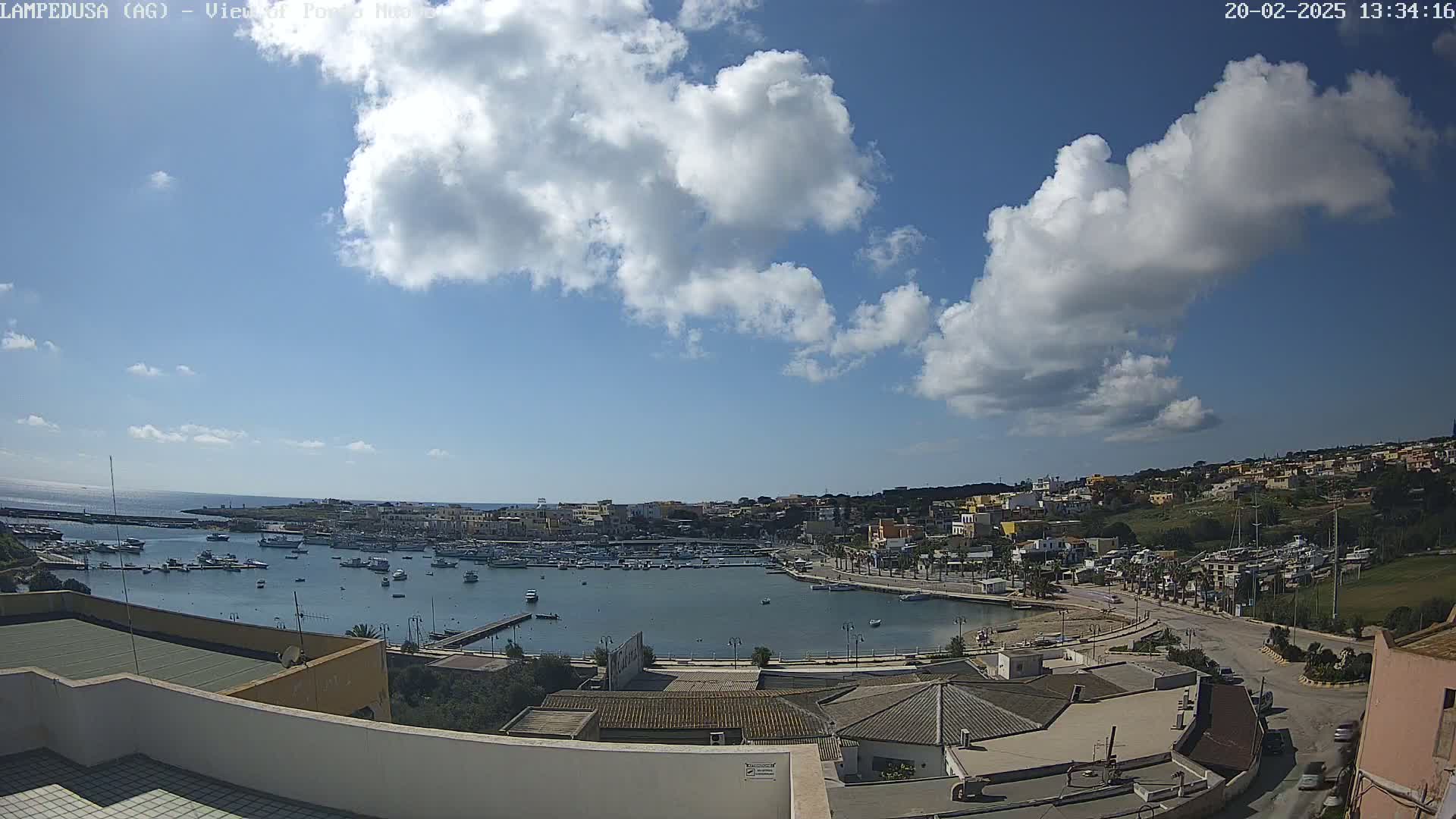 A partly sunny day reveals a harbor filled with boats, surrounded by a town nestled against a hillside, all under a sky dotted with fluffy cumulus clouds.