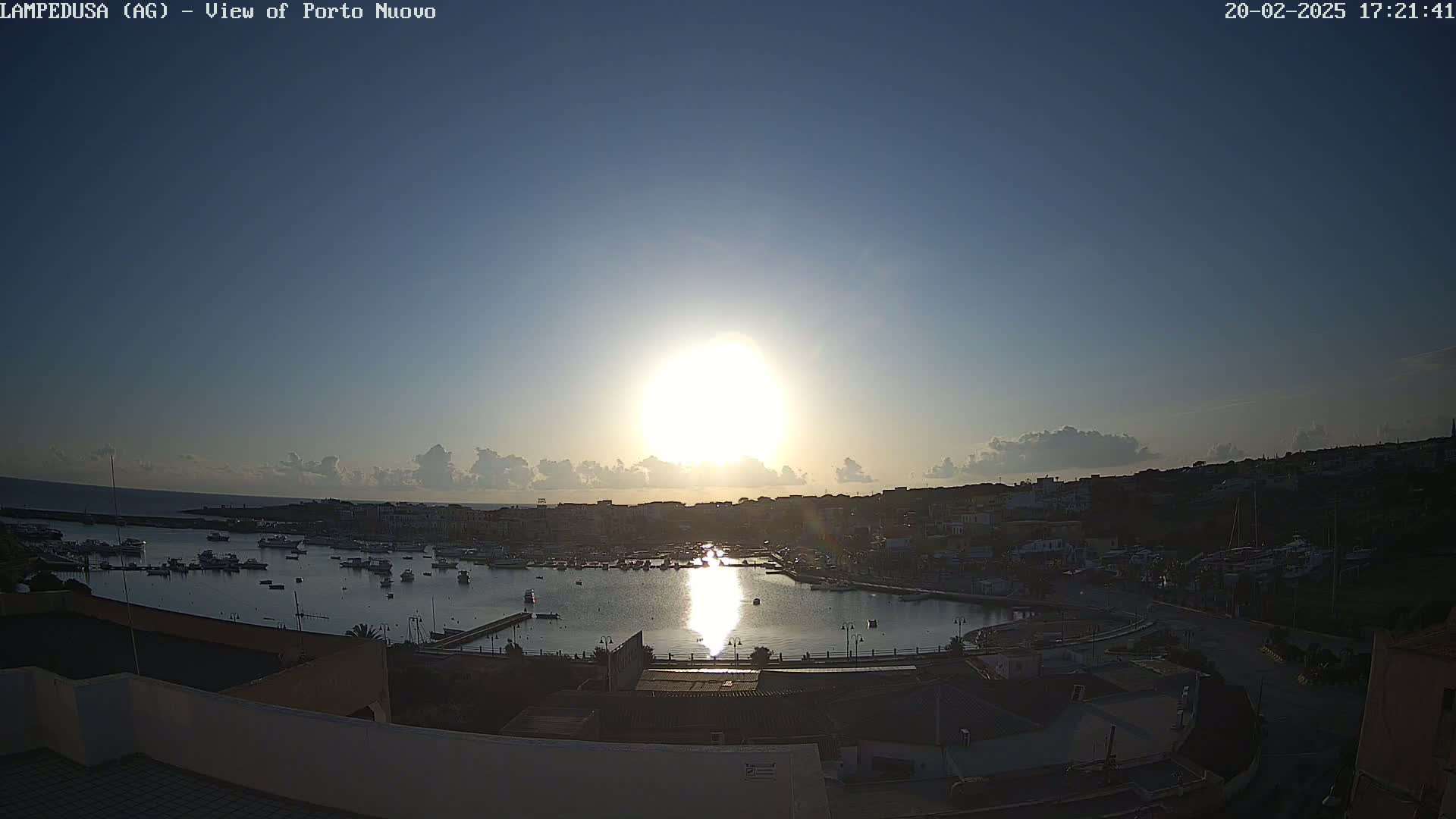 A bright sun shines over a harbor filled with boats, with a town situated on a hill overlooking the water under mostly clear skies.
