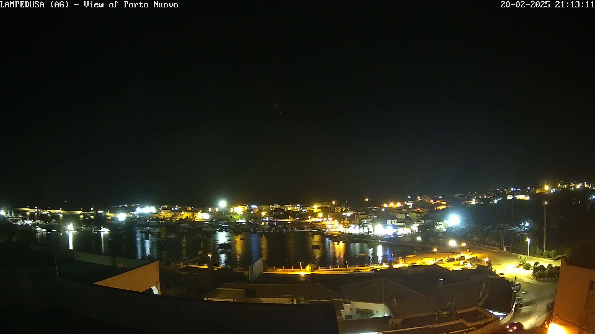 A nighttime, clear-weather view of a harbor town with boats in the water, illuminated buildings lining the shore, and surrounding hills.