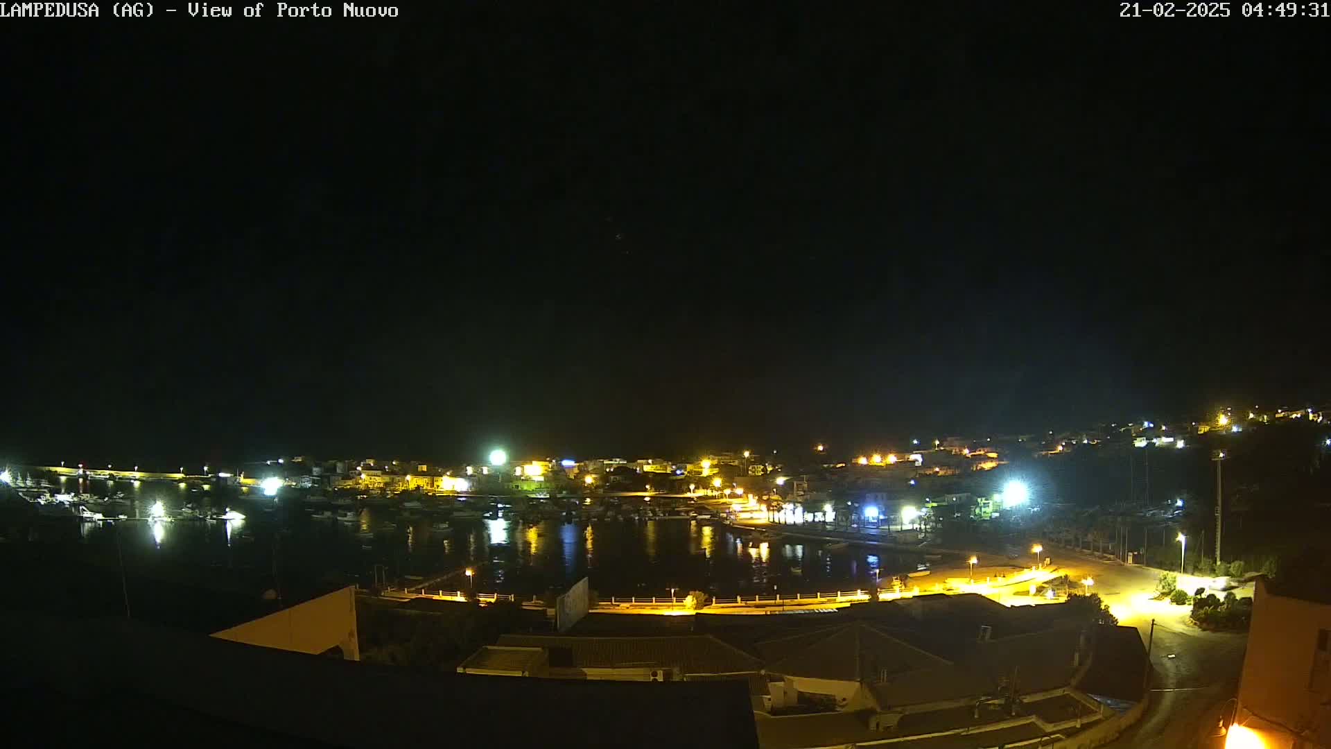 A nighttime view of a harbor town, with boats in the water and buildings illuminated along the shoreline under a clear, dark sky.